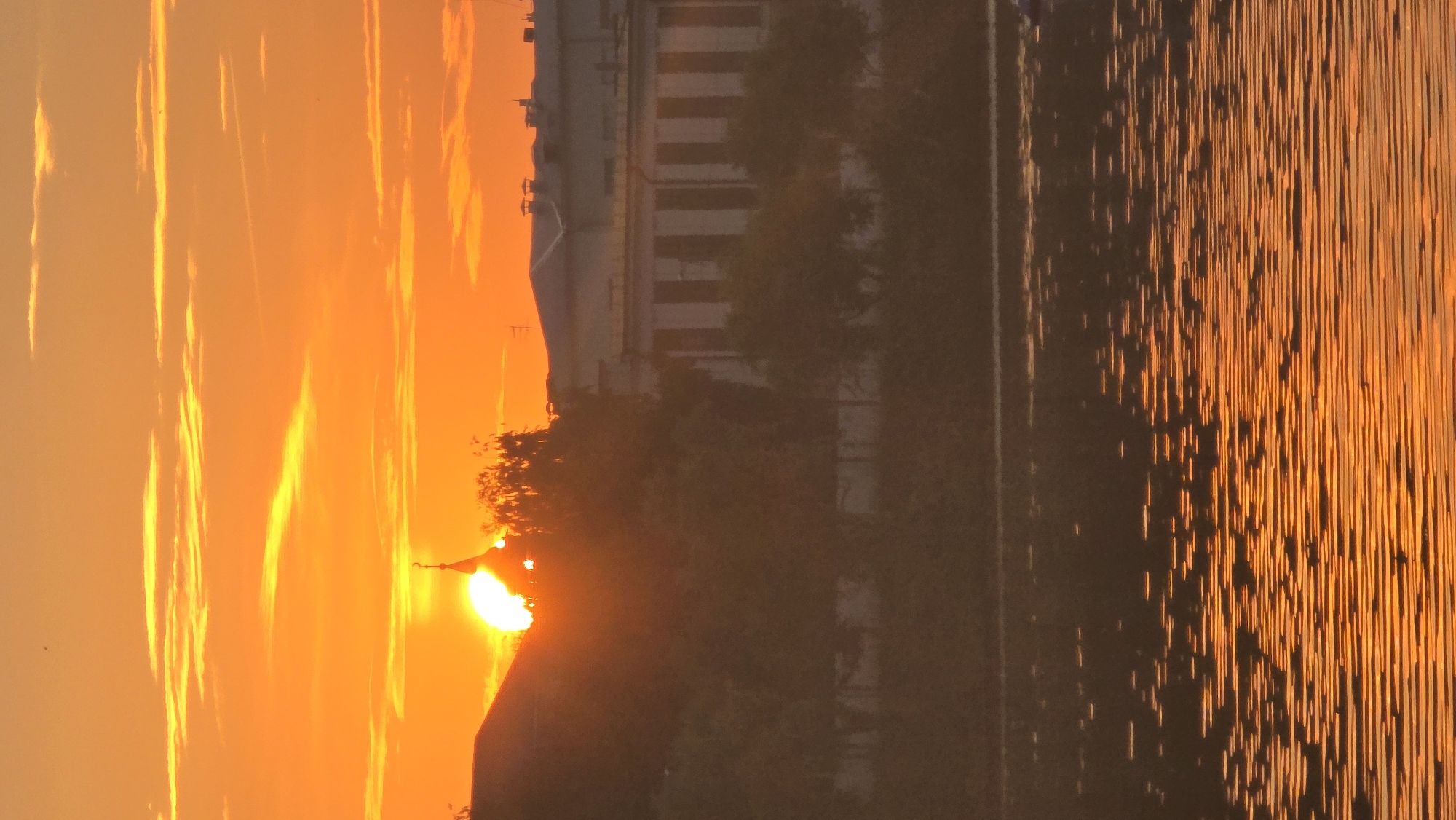 The image captures a vibrant sunset over a body of water, likely a river or lake, in Kazan, Russia. The sky is a brilliant orange, filled with streaks of golden clouds. The sun, a bright disc, is partially obscured by the silhouette of buildings and trees, casting a warm glow over the scene. In the foreground, the water reflects the sunset, creating shimmering patterns of orange and gold. A long, low-lying building with a distinctive facade stands in the middle ground, its details softened by the dusky light. The overall impression is one of serene beauty, characteristic of a sunset over a Russian city.