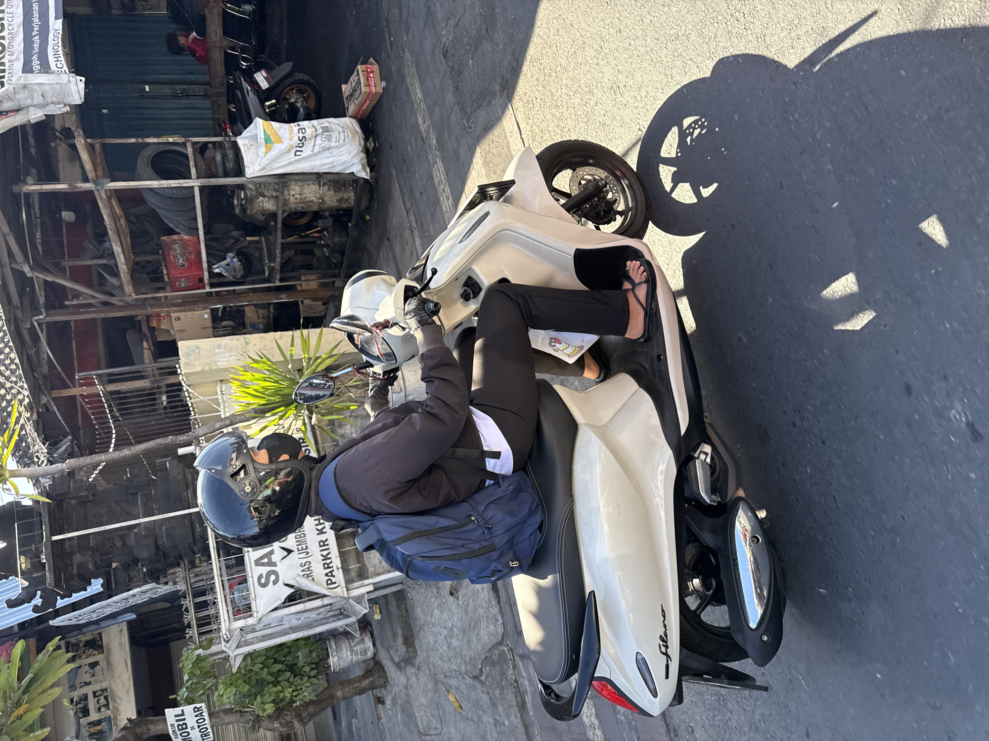 A person wearing a black helmet and a blue backpack rides a cream-colored scooter on a street in Denpasar, Indonesia. The person is wearing a dark jacket, dark pants, and black flip-flops. The scooter is a Yamaha Filano. In the background, there is a motorcycle repair shop with tires, tools, and parts. A sign on the shop reads "SARAS JEMBAR (PARKIR KH)". The sun is shining, casting long shadows on the road.