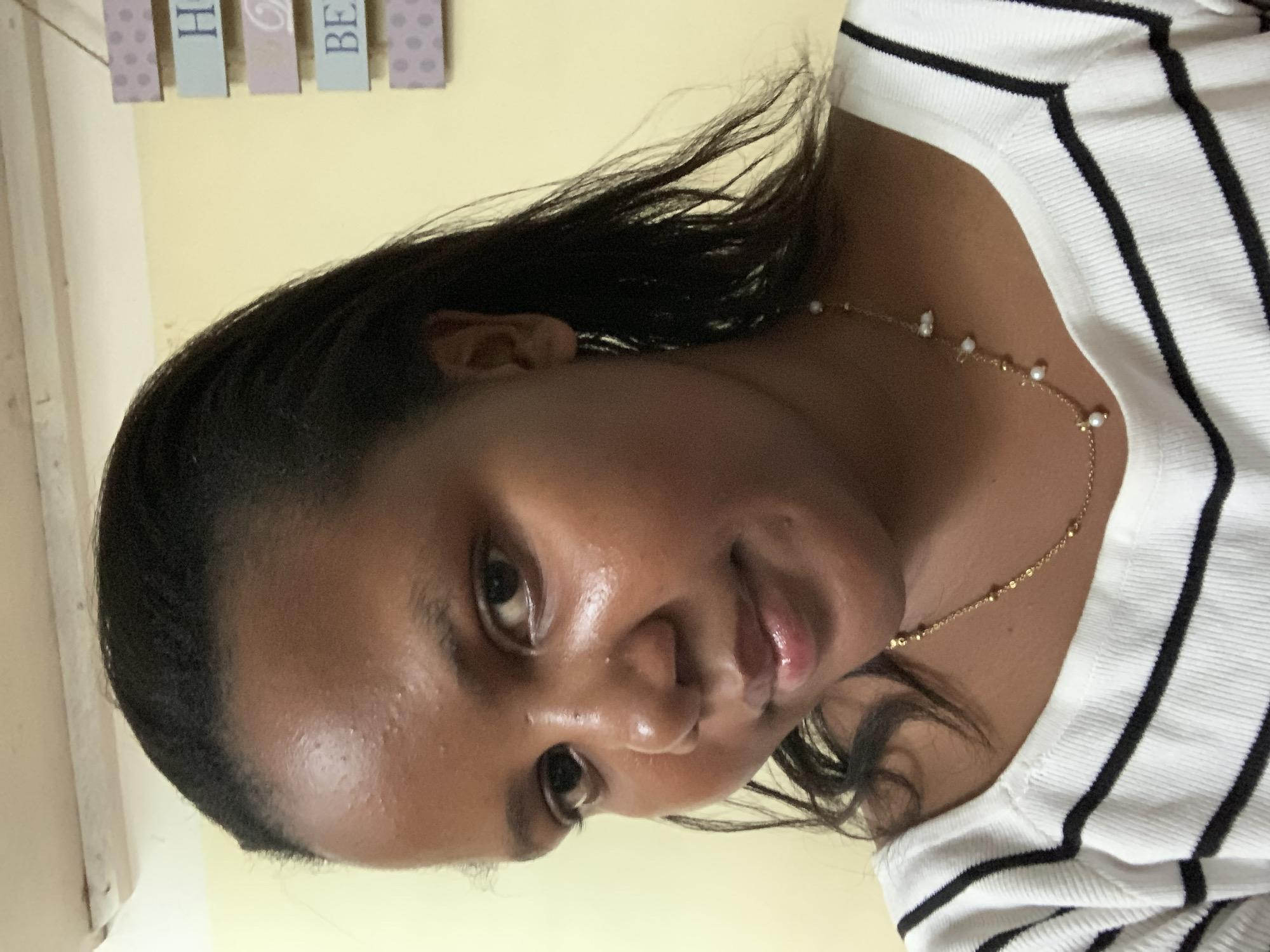 A close-up, slightly angled shot captures a woman with dark hair wearing a white and black striped shirt and a delicate gold necklace with small pearl-like accents. The background is a plain yellow wall with decorative plaques hanging above. No information suggests this image was taken in Dar es-Salaam, Tanzania.