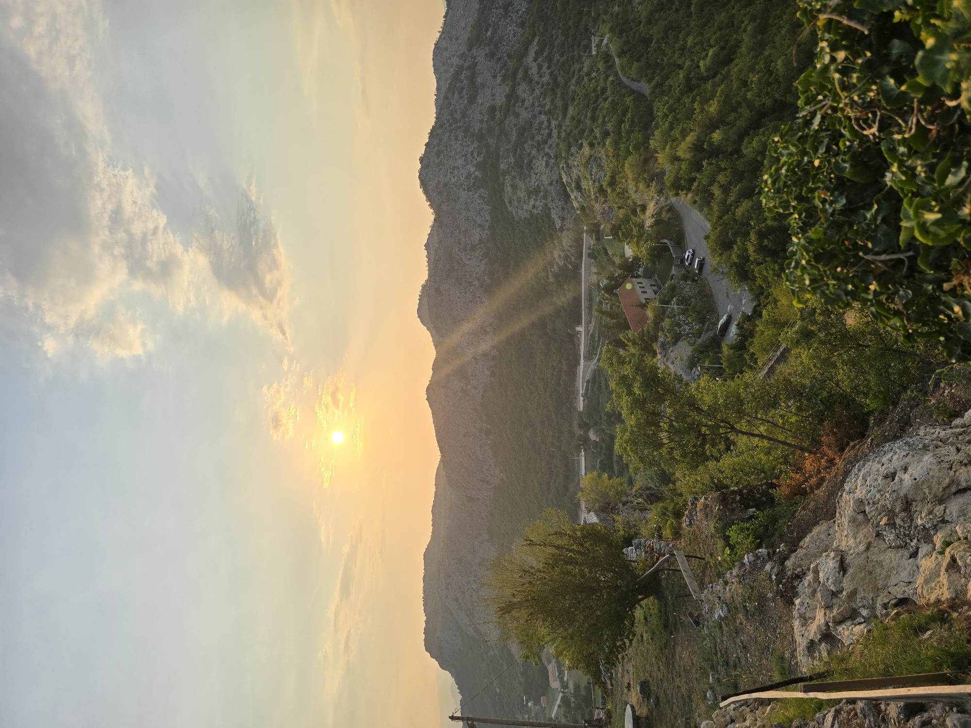 This image captures a scenic view of the landscape surrounding Medun, Montenegro, at sunset. The sun, low in the sky and casting a warm, golden light, is partially obscured by scattered clouds. Sunbeams radiate downwards, illuminating the rugged, forested hills that dominate the background. A winding road snakes through the greenery, leading to a cluster of buildings, one of which is a reddish-tiled house. In the foreground, the rocky terrain is covered with vegetation, including trees and bushes. A wooden fence lines the edge of the viewpoint, and a stone bench is visible to the left. The overall impression is one of tranquility and natural beauty, characteristic of the Montenegrin countryside.