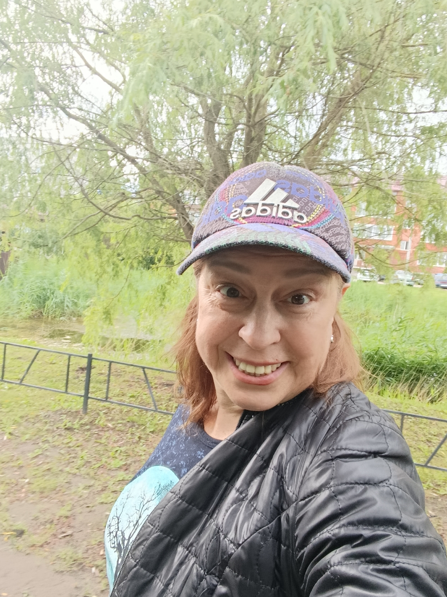 A woman with shoulder-length auburn hair and light skin is captured in a selfie outdoors. She is wearing a black, quilted jacket over a dark blue shirt with a light blue tree graphic. On her head, she sports a multi-colored baseball cap with the Adidas logo prominently displayed. The woman is smiling broadly, her teeth visible, and her eyes are looking directly at the camera.
The background reveals a natural scene with lush green foliage and a large tree with delicate branches and light green leaves. Beyond the greenery, a brick building with multiple windows is visible, suggesting an urban or suburban setting. A black metal fence runs horizontally across the lower part of the image. The ground appears to be damp and covered with dirt and some patchy grass. The overall lighting suggests an overcast day, perhaps late morning or early afternoon, with diffused natural light. The weather appears to be slightly damp or recently rained, given the appearance of the ground. The text "adidas" is visible on the woman's cap. The location context provided is Verea, Russia.