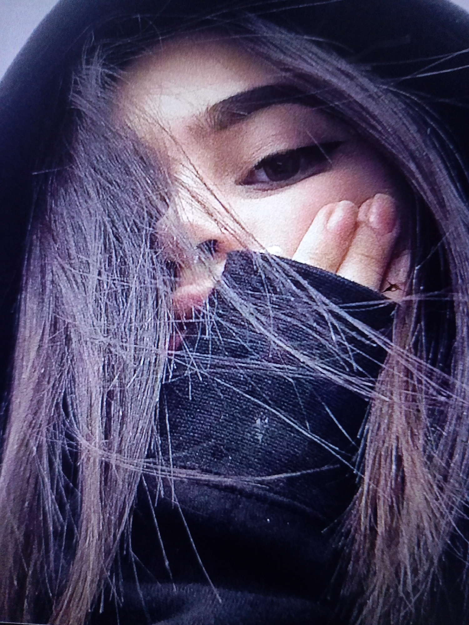 The image shows a close-up of a person's face, partially obscured by their hair and a dark fabric, possibly a hoodie or scarf. The person's eyes are visible, with one eye looking directly at the camera. Their hand is raised, with fingers touching their cheek. The hair is dark and appears to be long, falling across the face. The background is dark and out of focus, providing no discernible environmental details. The image does not contain any information about the city of Dobra Krynytsia, Ukraine.
