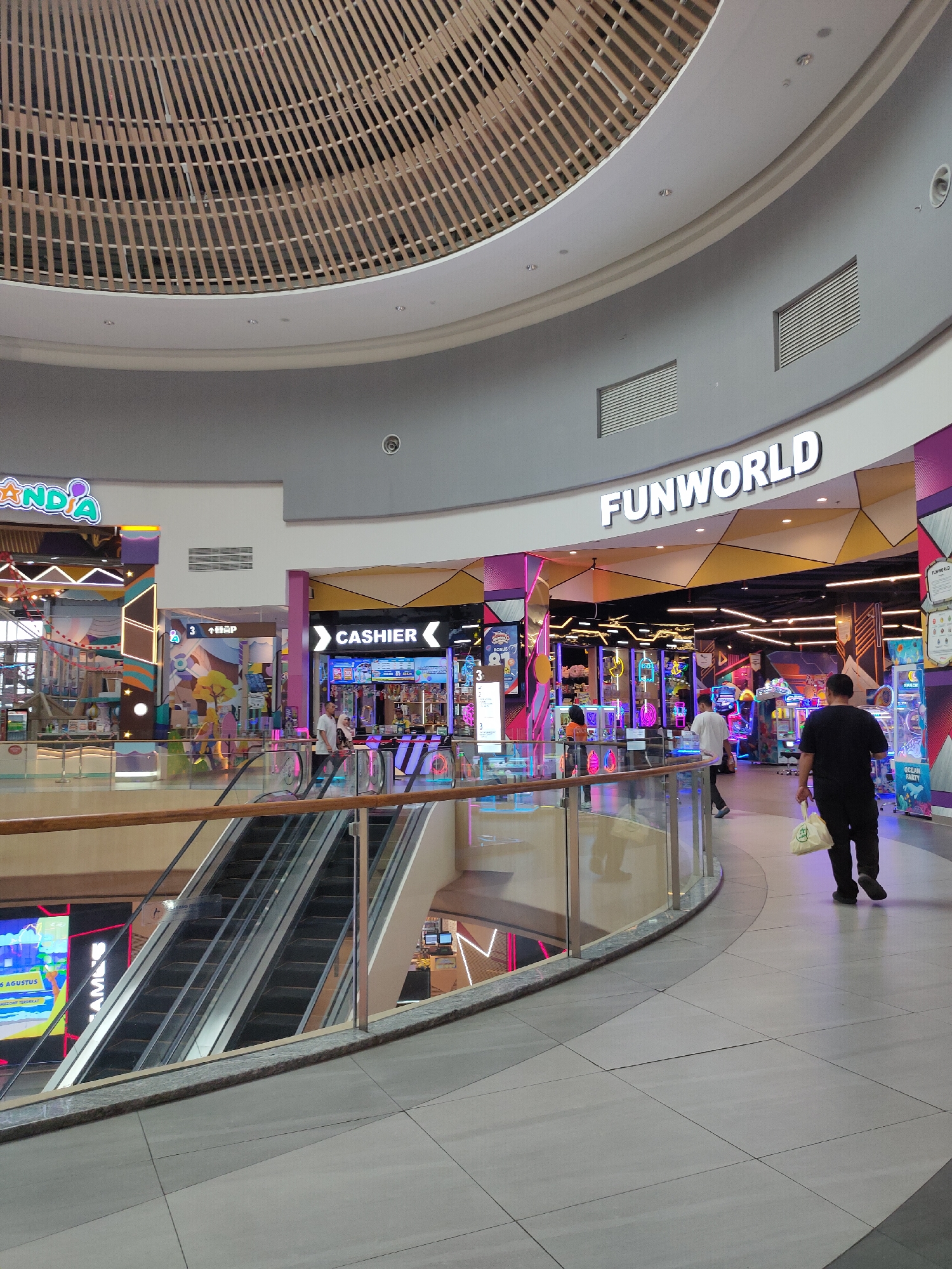 The image shows the interior of a shopping mall, likely located in Lambangjaya, Indonesia. The scene is brightly lit with a mix of ambient light and neon lighting from various storefronts. In the foreground, a curved hallway with a tiled floor leads the eye towards the right. A person in a black t-shirt and black pants is walking away from the camera, carrying a light-colored bag in their right hand. To the left, escalators ascend between floors, with a glass railing overlooking the level below. The upper level of the mall features a large sign that reads "FUNWORLD" in white, illuminated letters, indicating an amusement or entertainment center. Adjacent to "FUNWORLD," another sign reads "CANDIA" and appears to be another entertainment venue. Several other storefronts with colorful displays are visible in the background, contributing to the vibrant atmosphere. The ceiling is a striking feature, with a large, circular wooden latticework design.