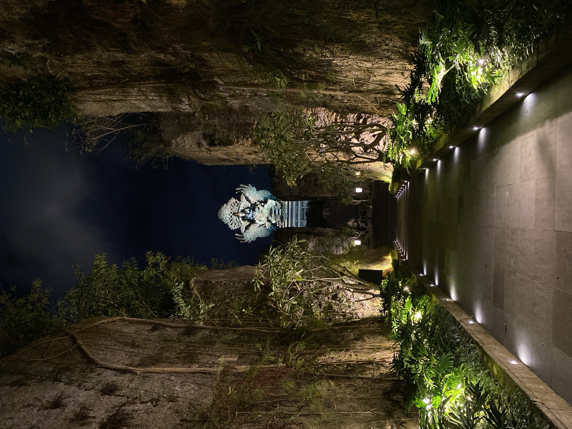 In Kuta Selatan, Indonesia, at night, a pathway leads towards a large statue of Garuda Wisnu Kencana, illuminated by spotlights and ambient lighting. The pathway is flanked by tall, rough-hewn rock walls covered in vines and lush green foliage. Small, square lights are embedded along the edge of the pathway, casting pools of light onto the stone pavement and the surrounding plants. Trees with leafy branches grow in front of the rock walls, their foliage also lit from below. In the distance, the grand statue, depicting the Hindu deity Vishnu riding the mythical bird Garuda, is prominently visible, bathed in a bright, cool light. The sky above is dark, with hints of clouds. A few individuals can be seen in the far distance, near the base of the statue, suggesting this is a public area. The overall atmosphere is dramatic and somewhat mystical due to the nighttime lighting and the imposing scale of the monument.