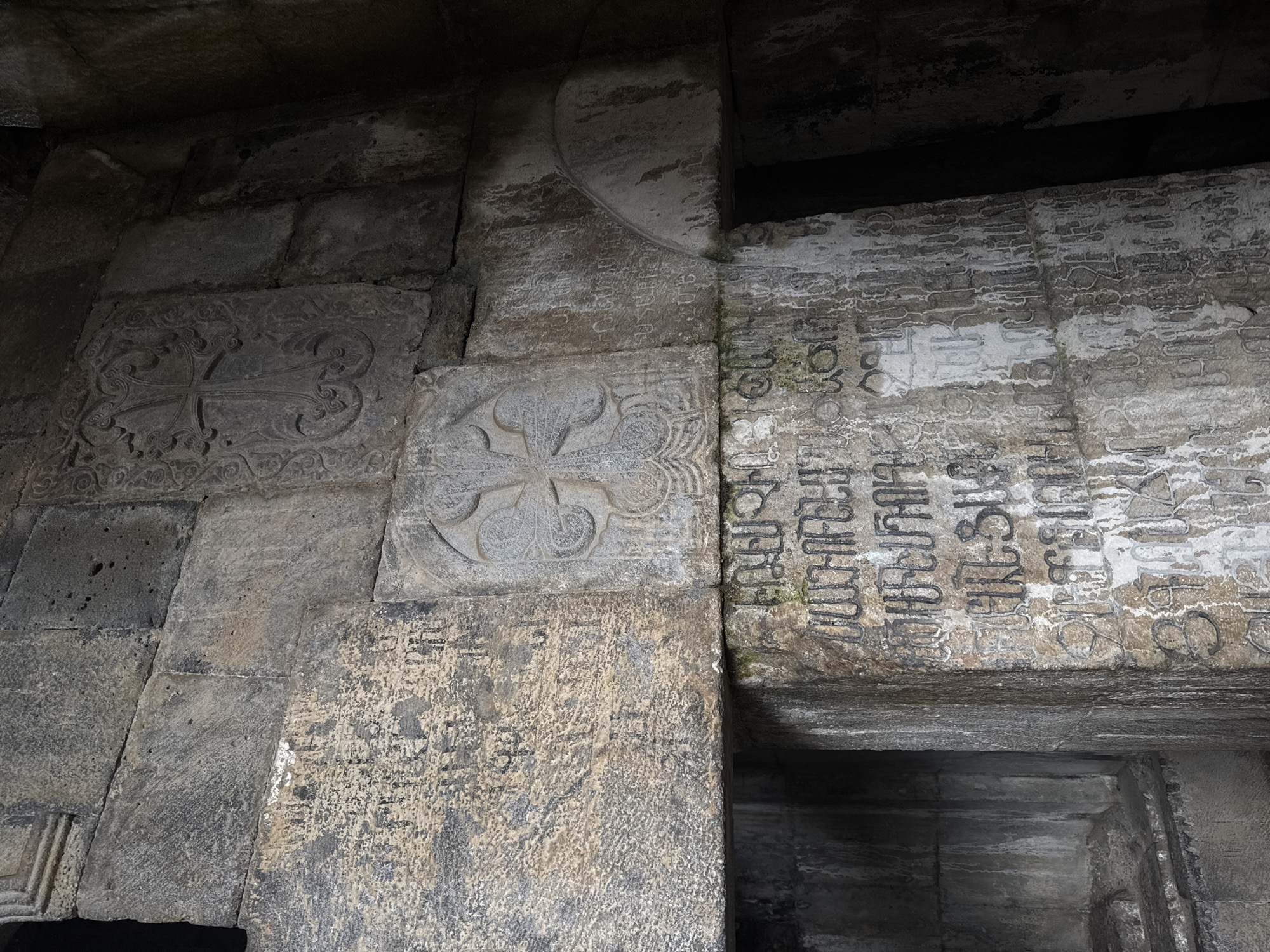The image displays a close-up view of weathered stone carvings and inscriptions within an ancient structure. The stonework shows signs of age and erosion, with some areas appearing rough and pitted, while others exhibit clearer carved details. Prominently featured are two carved crosses. The first, located in the upper right quadrant of the image, is a relief carving of a cross with ornate swirling patterns surrounding it. Below this, in the center of the image, is another carved cross, this one appearing more abstract with petal-like shapes emanating from its center.
The lower portion of the image is dominated by a dense collection of inscriptions carved into the stone. These inscriptions are in an unfamiliar script, appearing as a series of symbols and characters. Some of these inscriptions are faded or partially obscured by the rough texture of the stone and possible water staining. The overall environment appears to be an interior or covered space within a historic building, likely a monastery given the religious iconography. The lighting is dim, casting shadows and highlighting the textures of the stone.
The location is identified as Haghpat, Armenia. This specific site is part of the Haghpat Monastery, a UNESCO World Heritage Site known for its rich history and significant examples of Armenian medieval architecture and religious art. The carvings and inscriptions likely represent religious texts, historical records, or dedications from various periods of the monastery's existence. No people are visible in the image. The focus is entirely on the architectural details and the inscribed surfaces.
