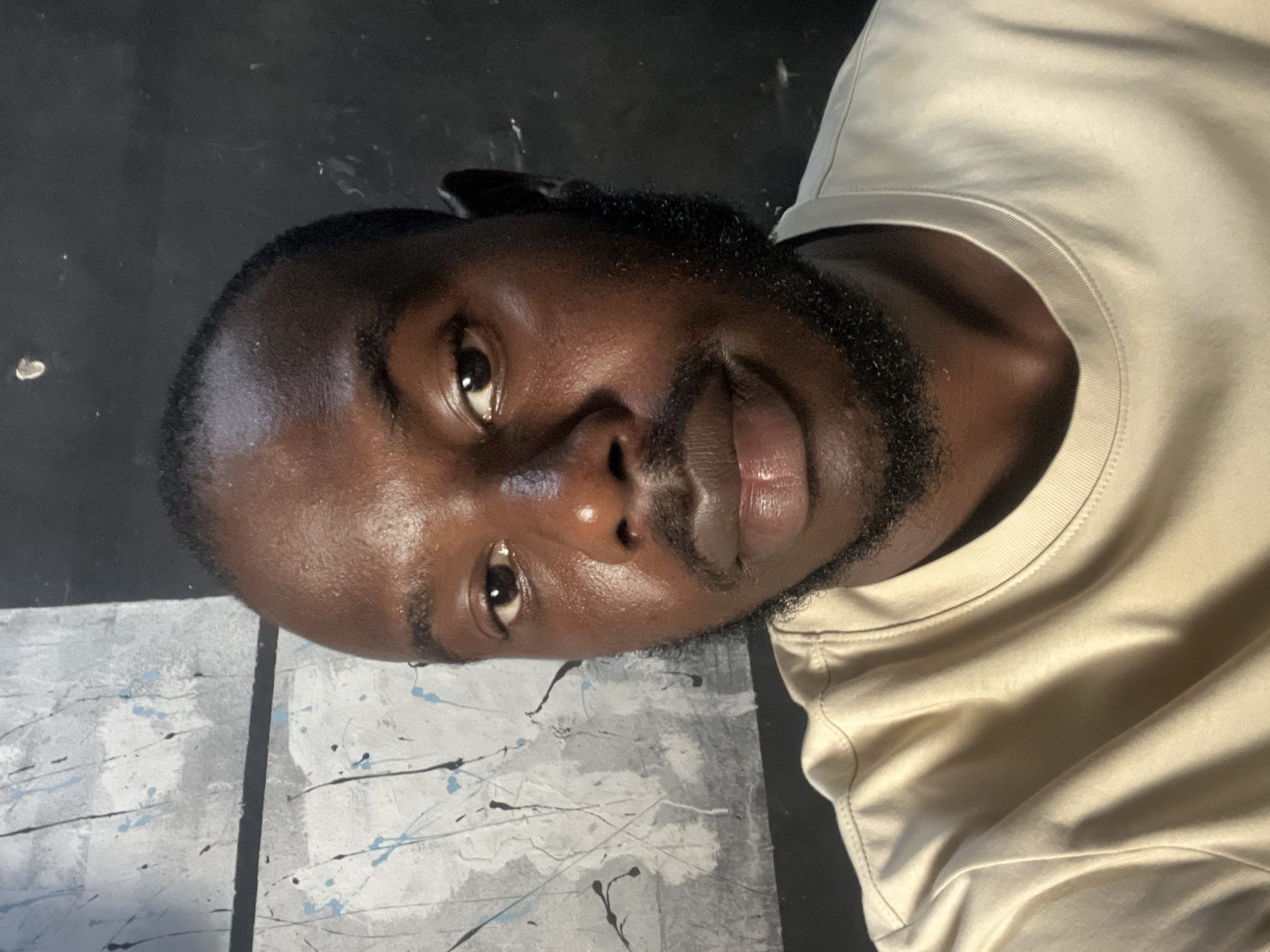 The image is a close-up, waist-up portrait of a Black man. He has short dark hair, a beard, and is wearing a light beige t-shirt. The background is divided vertically into two sections: the left side features a grey and white abstract pattern with black and light blue splatters, and the right side is a dark, slightly textured surface, possibly a wall. The lighting appears to be coming from the front, casting subtle highlights on his skin and t-shirt. No specific location in Jos, Nigeria, can be identified from the image.