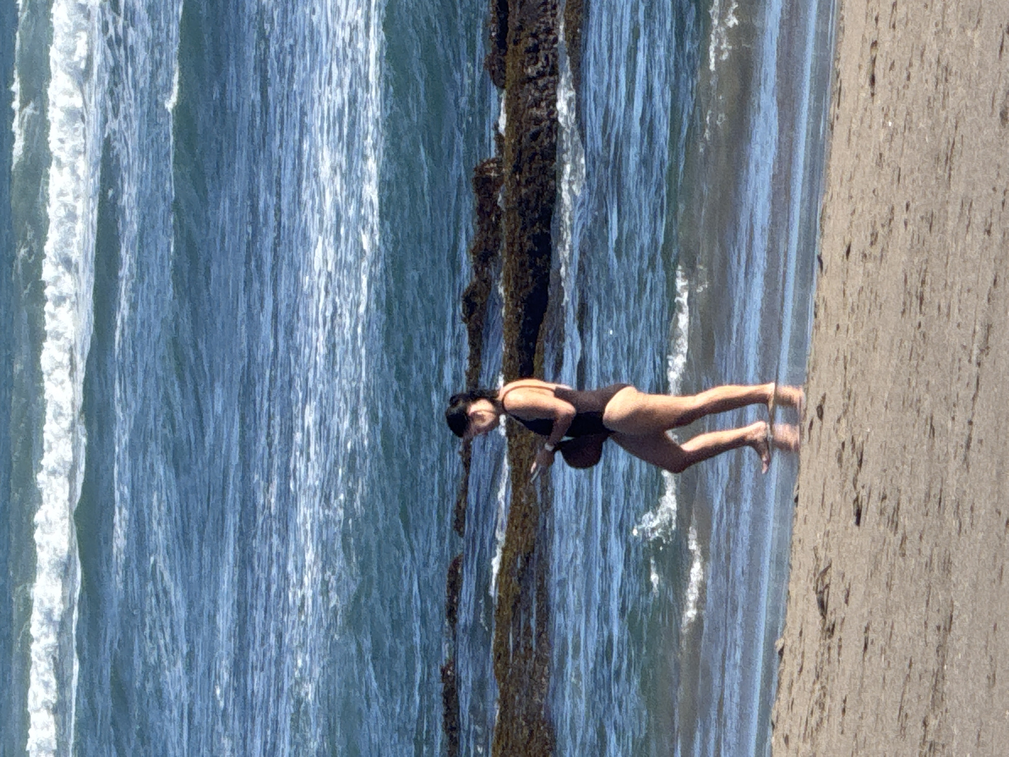 A woman in a dark one-piece bathing suit stands on a beach in North Kuta, Indonesia, next to the ocean. The beach is tan and the water is blue with whitecaps. The woman is holding a dark object in her right hand. An algae-covered seawall is between the woman and the ocean.