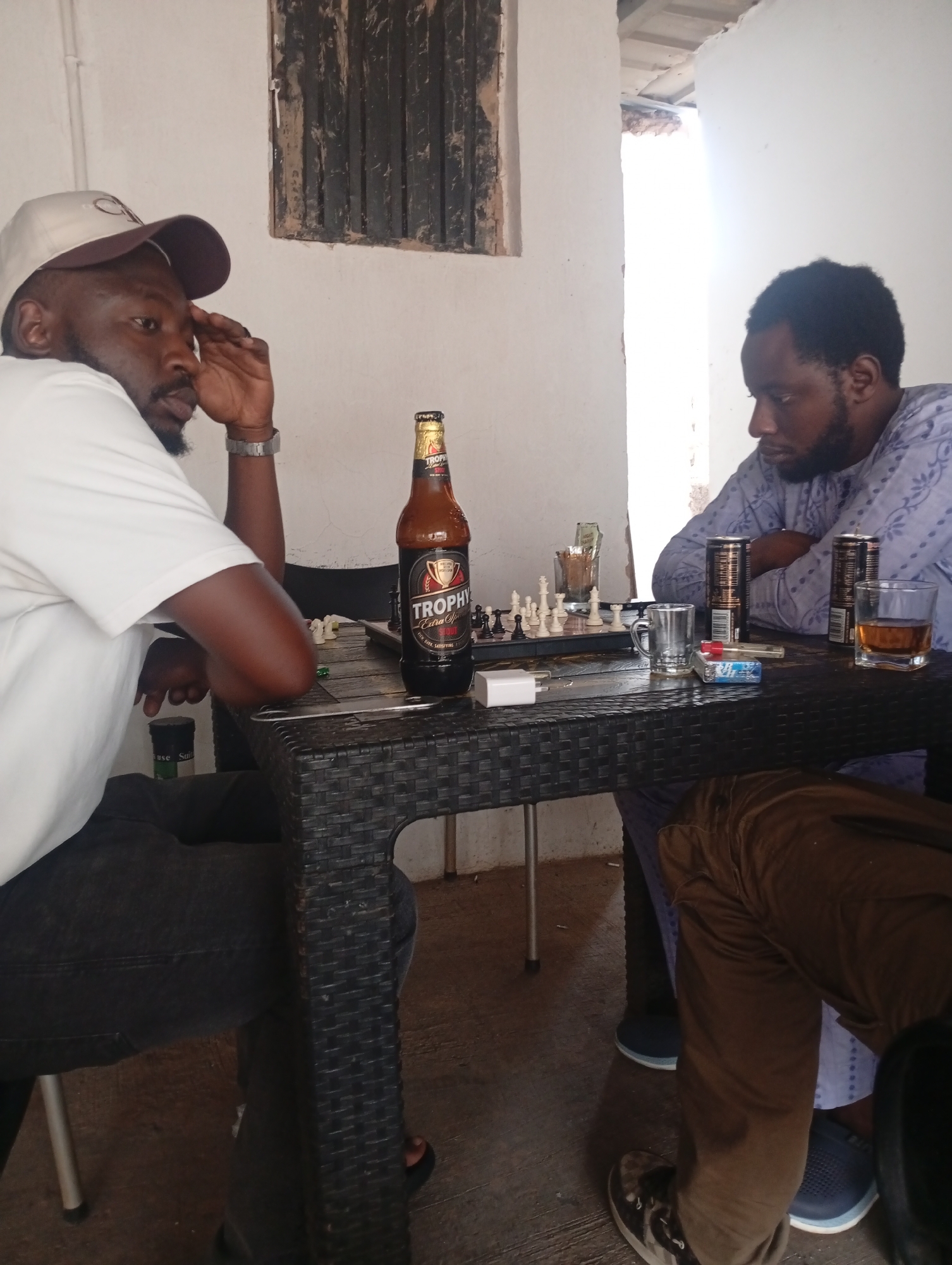Two men are seated at a dark, possibly wicker-style table, engaged in a game of chess in a casual indoor or covered outdoor setting. The man on the left wears a white t-shirt, dark trousers, and a light-colored baseball cap with "TROPHY" visible on it. He has a beard and is resting his head on his hand, looking intently at the chess board, suggesting deep thought or focus. A silver-toned watch adorns his left wrist.
The man on the right, also bearded, is dressed in a light blue, patterned long-sleeved top, possibly a traditional garment. He wears brown trousers and blue sandals, and is leaning forward, observing the chess pieces.
Between the two men, a prominent brown bottle of "Trophy Extra Stout" beer stands on the table. A chess board is set up, with white pieces clearly visible on their starting squares. Surrounding the game, there are several other items: two dark-colored beverage cans, a clear glass holding an amber liquid, a pack of cigarettes and a lighter, a clear glass mug, and a white phone charger. A glass containing what appears to be currency is visible behind the chess board, and a small salt shaker is partially seen under the table.
The background features light-colored walls. On the left wall, there is a dark, weathered wooden window frame or shutter structure. To the right, a bright opening suggests an entrance or large window, providing ample natural light and indicating it is daytime. The floor is a concrete or tiled surface. The scene portrays a relaxed social interaction, likely centered around the game of chess and drinks.