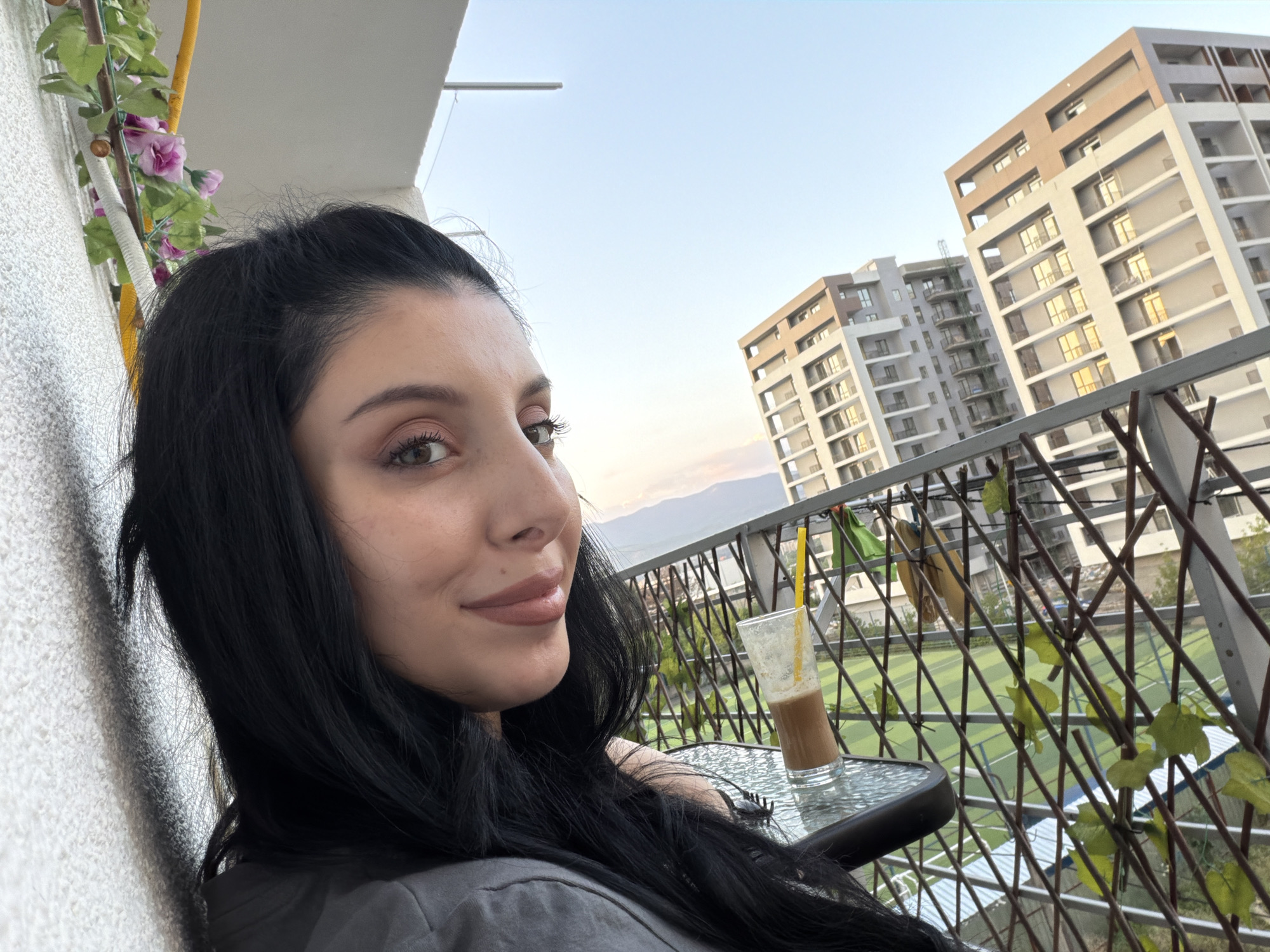 In Tbilisi, Georgia, a young woman with long, dark hair is captured in a selfie on a balcony. She is seated near a small glass table, on which rests a glass containing a brown beverage with a yellow straw. The woman is looking towards the camera with a slight smile. Behind her, the scene opens up to reveal modern apartment buildings and a clear sky. A balcony railing with decorative vines stretches across the foreground, partially obscuring a view of a green field, possibly a sports field, below. The overall setting suggests an urban environment with residential buildings and recreational space.
