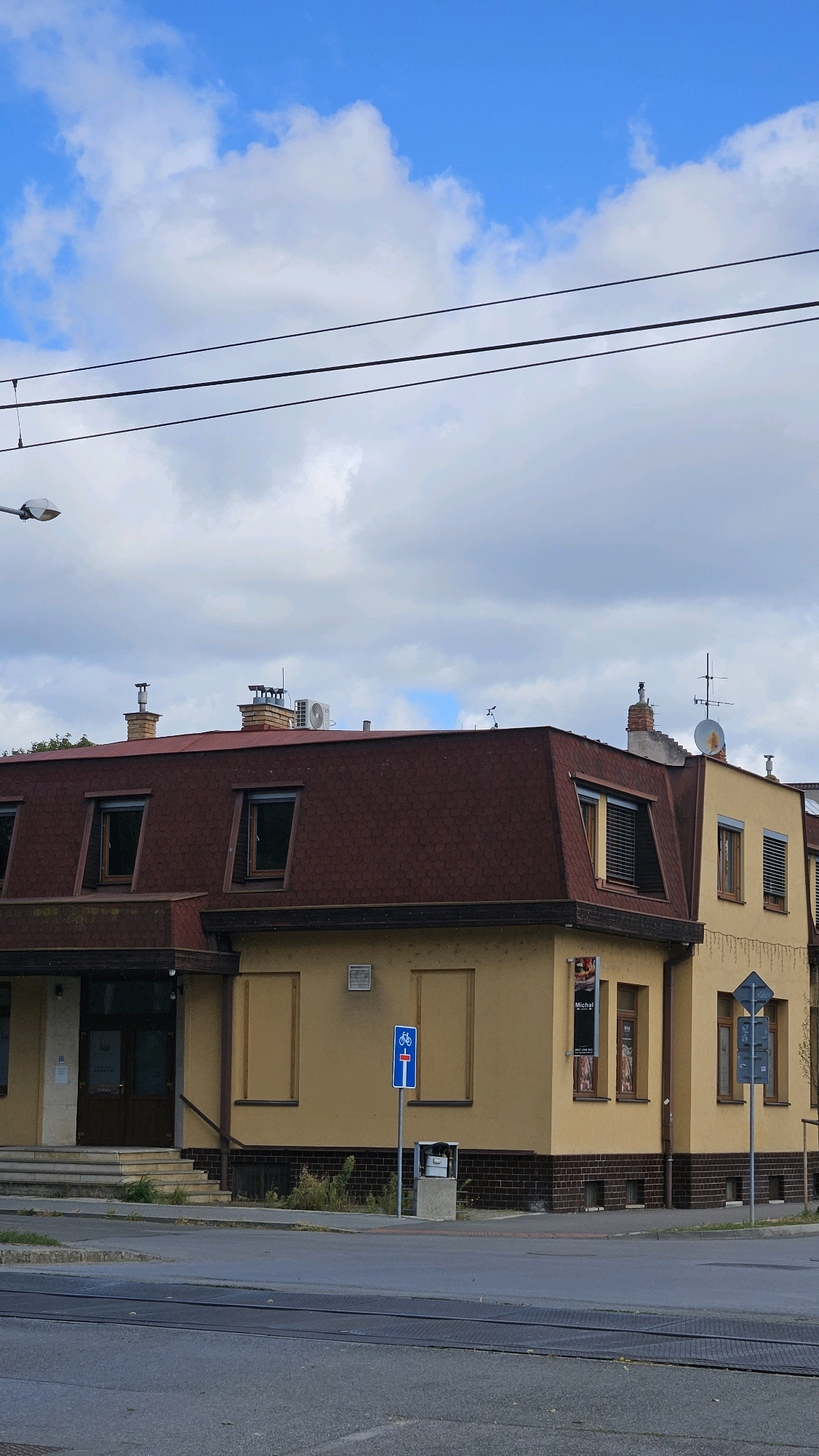 The image shows a yellow building with a dark red, mansard-style roof. The building has several windows, some with dark shutters. There are also two chimneys and an air conditioning unit on the roof. A street sign with a bicycle symbol and a blue rectangle is visible on the right side of the building. The building appears to be located on a street with tram tracks in the foreground. In the background, there is a blue sky with white clouds. The image is taken from a street level perspective. Based on the street sign and the architectural style, this building is located in Prešov, Slovakia, specifically at the intersection of Budovateľská and Štefánikova streets. The building is likely a commercial establishment or residential building.