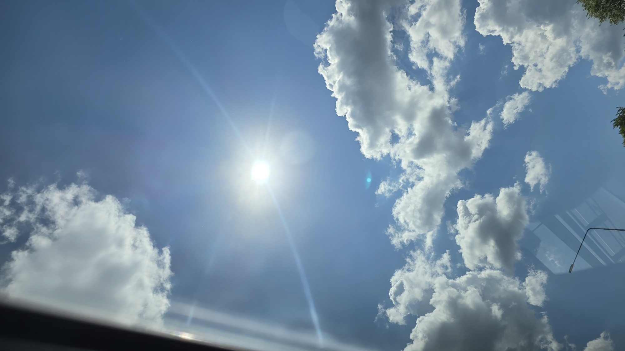 A vertical, full-frame photograph captures a bright, sunny sky with scattered white clouds. The sun is prominently positioned in the upper center of the frame, emitting strong rays that create lens flare and a visible blue light artifact. A dark, vertical element, possibly a window frame or obstruction, is visible on the left edge of the image. In the lower portion of the frame, a street lamp with a curved arm is visible, and reflections of what appear to be windows or a ceiling are superimposed on the sky. Some green foliage can be seen at the bottom right corner, suggesting an outdoor environment. The scene is consistent with a view from a vehicle or indoor location looking outwards on a clear day. The location is specified as Tolbazy, Russia.