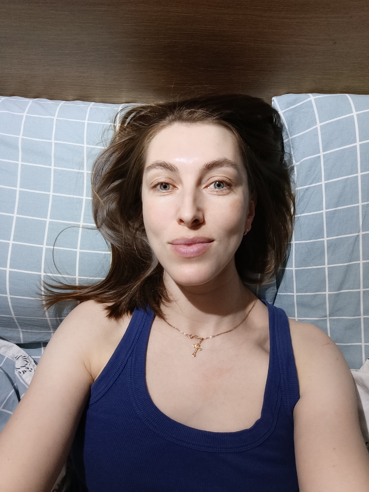 A woman is lying on a bed. The woman is white, with brown hair and blue eyes. She is wearing a blue tank top and a gold necklace with a cross pendant. The bed has a blue and white checkered pillow. Behind the pillow is a wooden headboard. The location is presumed to be indoors, but there is no information available to determine if it is in Chaikovsky, Russia.