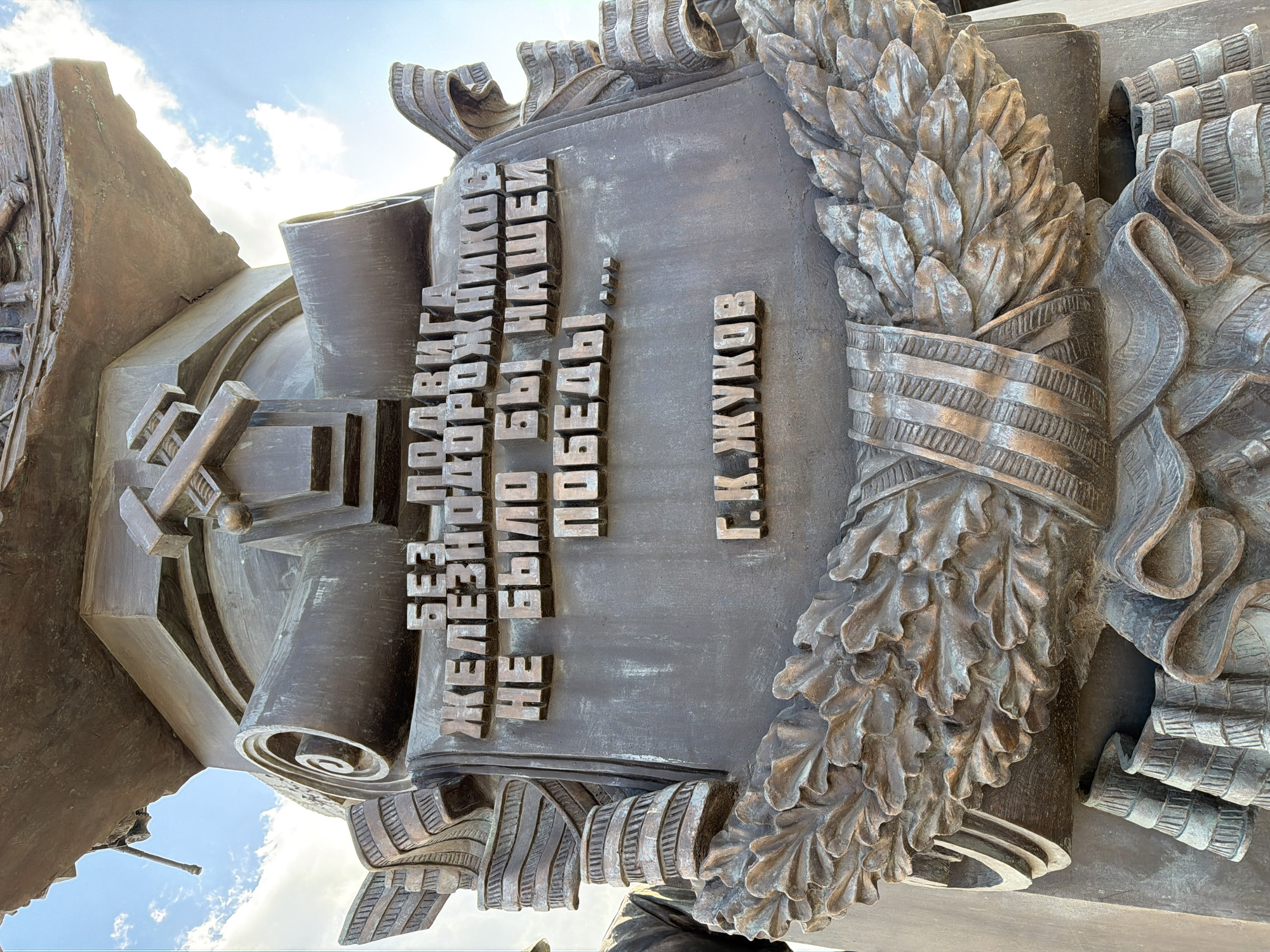 The image displays a detailed section of a large bronze monument, likely commemorative, situated outdoors under a partly cloudy sky in Moscow, Russia. The central focus is a robust bronze plaque featuring raised Cyrillic text. The main inscription reads: "БЕЗ ПОДВИГА ЖЕЛЕЗНОДОРОЖНИКОВ НЕ БЫЛО БЫ НАШЕЙ ПОБЕДЫ...", which translates to "Without the feat of railroad workers, there would be no our victory...". Below this, the text attributes the quote to "Г.К. ЖУКОВ" (G.K. Zhukov), referring to Marshal Georgy Konstantinovich Zhukov.
The plaque is embellished with elaborate bronze decorative elements. To the right, a highly detailed laurel wreath, symbolizing victory and honor, is interwoven with a textured ribbon. Surrounding the inscription are additional stylized bronze ribbons or banners with striped patterns, adding to the monument's grand scale. The bronze material exhibits natural patination, with subtle hints of green and blue, characteristic of weathered metal.
In the background and surrounding areas, abstract, geometric bronze forms are visible, possibly representing industrial components or fortified structures, which complement the monument's theme of wartime effort. The scene is brightly lit by natural daylight, highlighting the texture and depth of the sculpture. The sky, visible in the upper sections, is blue with scattered white clouds, creating a peaceful backdrop for this historical tribute. The monument commemorates the vital contribution of railway workers to victory, as underscored by Marshal Zhukov's quote.