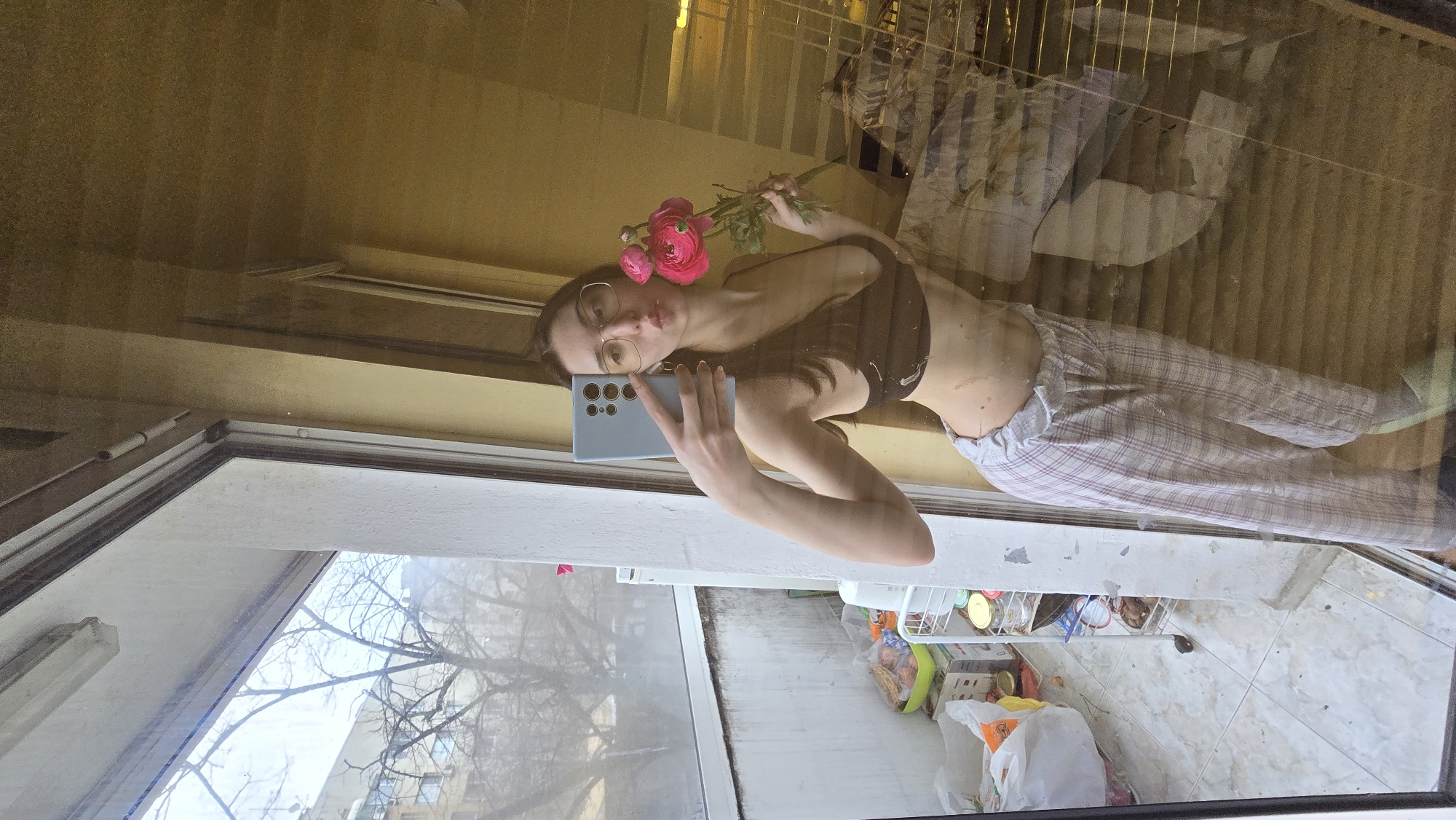 The image shows a woman reflected in a window, likely taking a selfie with a smartphone. She is wearing glasses, a dark bra, and plaid pajama pants. She is holding pink flowers near her face. The reflection shows an untidy room with various items on shelves and the floor. Through the window, trees and buildings are visible, which may place this location in the city of Stara Zagora, Bulgaria. The weather appears to be overcast.