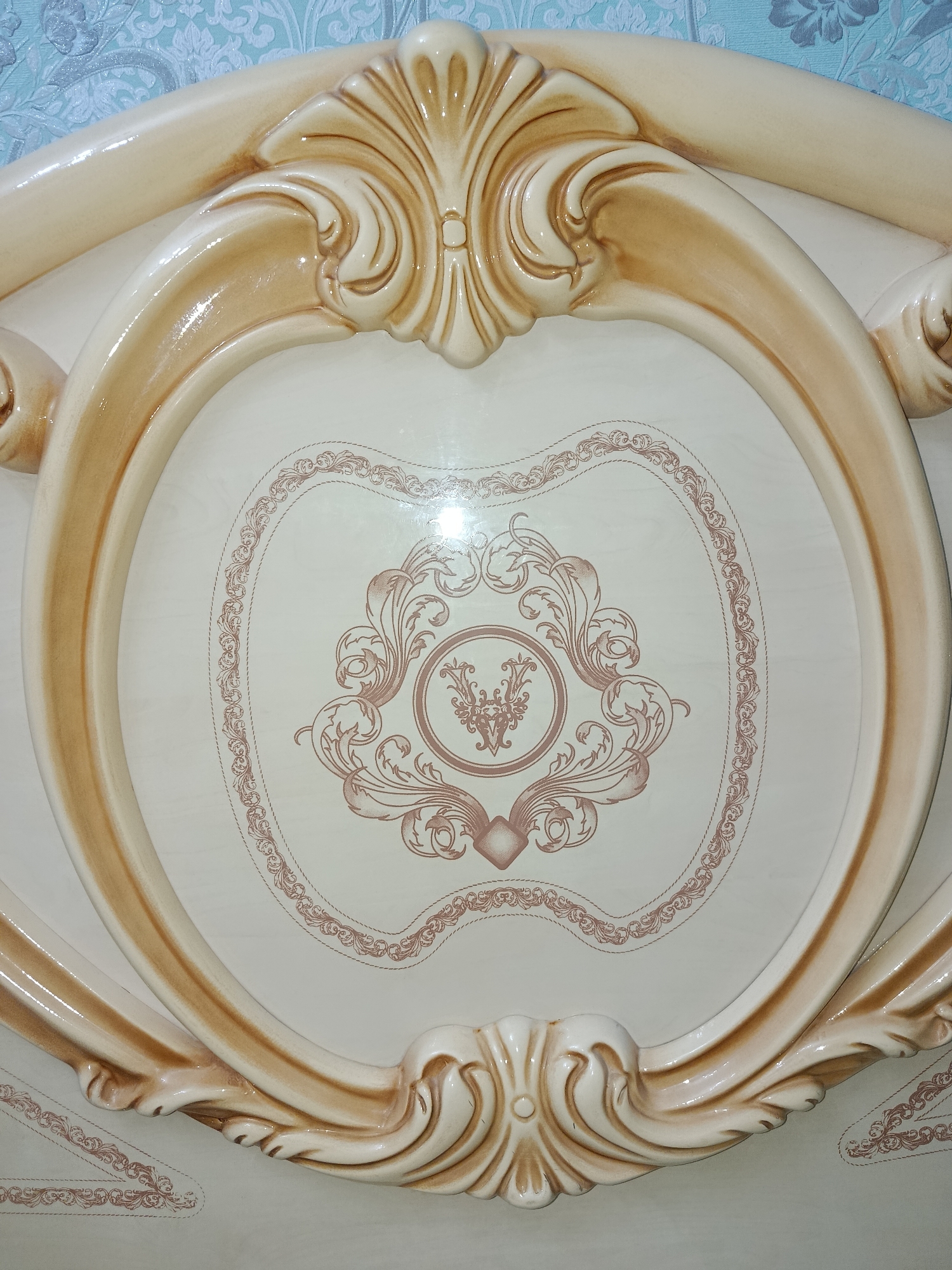 The image shows a close-up of a decorative headboard of a bed. The headboard is made of a light-colored material, likely wood or a composite, with a glossy finish. It features ornate carvings and a central oval panel. The carvings are in a creamy beige color with darker brown accents, emphasizing the details of the scrolling acanthus leaves and a central shell-like motif at the top and bottom. The oval panel is a pale, off-white color with a subtle wood-grain pattern. Within this panel, a detailed, intricate design in a muted rose or reddish-brown color is visible. This design consists of swirling vines, floral elements, and a circular emblem in the center, surrounded by a decorative border. The overall style is opulent and classical. The background of the headboard, visible at the top of the image, appears to be a patterned wallpaper with light blue and white tones. The lighting suggests an indoor setting, likely a bedroom. There is no visible text in the image. The image captures a static object and does not depict any activity or people. The context of Tashkent, Uzbekistan, does not provide any specific visual cues within the image itself but situates the object geographically.
