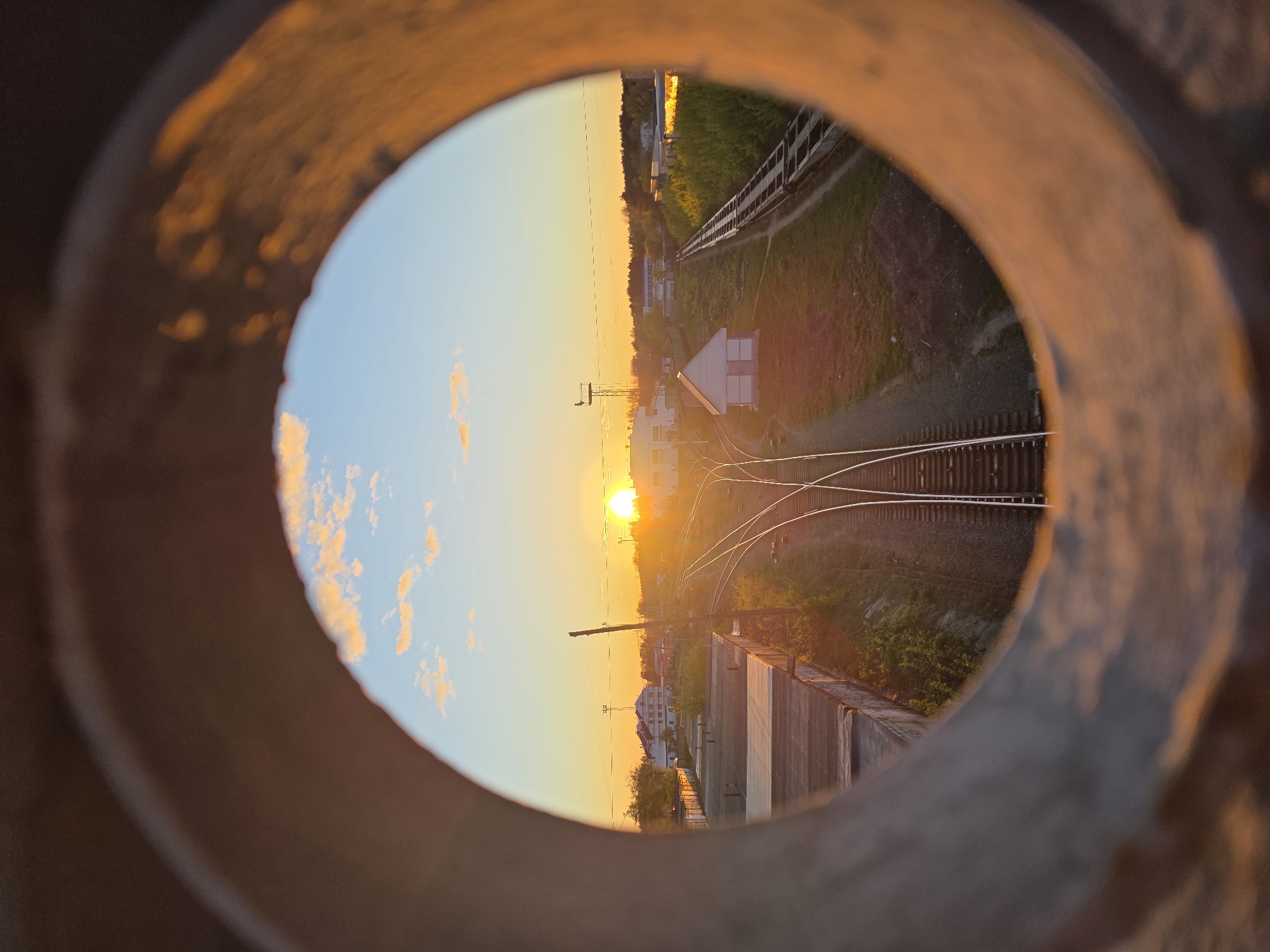 The image depicts a railway track at sunset, as seen through a circular opening, possibly a pipe or a hole in a structure. The sun is setting on the horizon, casting a warm orange glow across the sky and the landscape. The railway tracks diverge in the foreground, leading towards a small building and patches of vegetation. A train is visible on the railway in the distance. The circular opening appears to be made of metal, with an uneven, possibly rusty surface.
This scene is in the city of Chaikovsky, Russia.