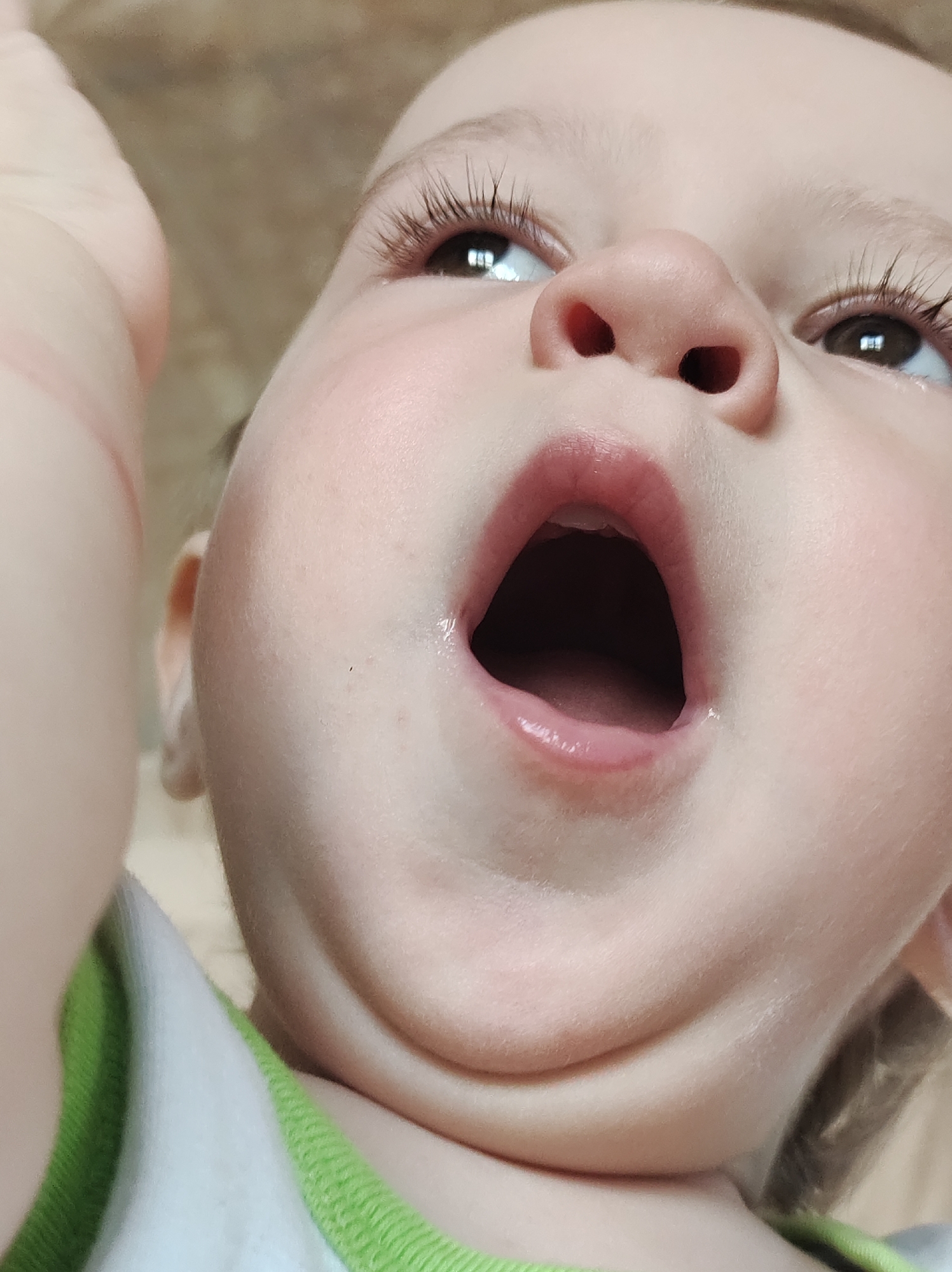 Close-up, low-angle view of a baby's face with mouth open. The baby has fair skin, brown eyes with long eyelashes, and a small nose. A hand is partially visible in the upper left corner. The baby is wearing a white shirt with a green trim. The background appears to be a blurred beige color. There is no indication in the image that it was taken in Фоки, Russia.