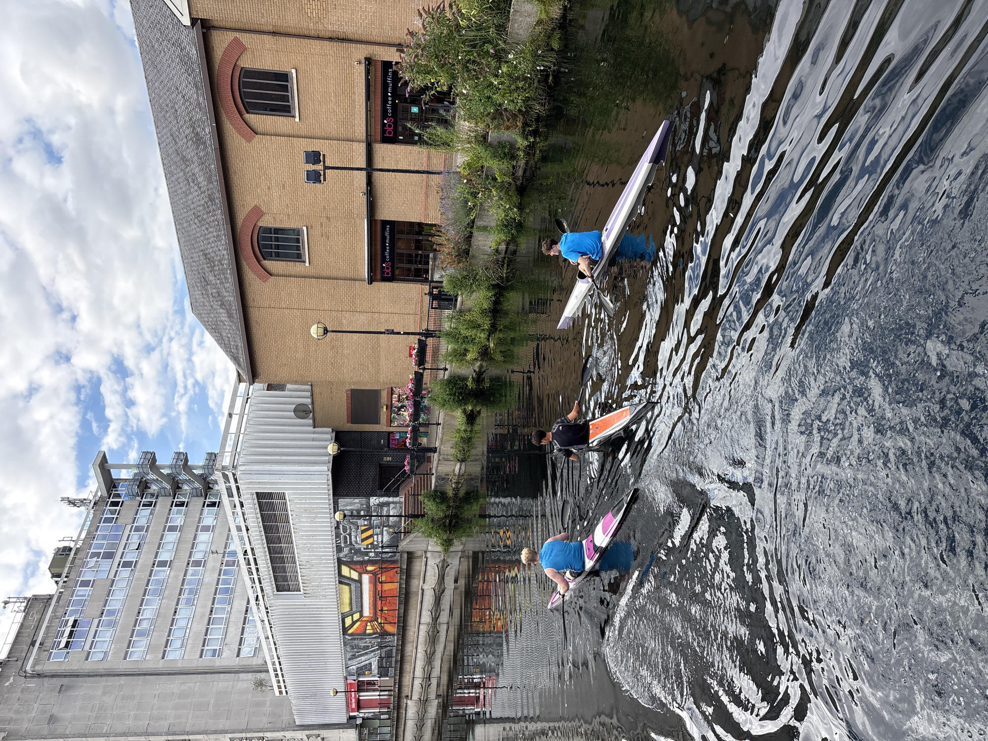 The image captures a scene of people kayaking on a canal. Three individuals are visible in kayaks. The first kayaker, wearing a blue shirt, is in the foreground, paddling with a black oar. The second kayaker, in a black shirt, is further back and slightly to the right, also paddling. The third kayaker, wearing a pink top, is in the distance and to the right, with their kayak pointing towards the viewer. The kayaks themselves are sleek and white with colored accents.
The setting appears to be an urban environment, with buildings lining one side of the canal. On the left, a tall, modern building with many windows is visible. Adjacent to it is a lower, brick building with arched windows. Above the brick building, there are signs for "bbs coffee & muffins." The water of the canal is choppy, reflecting the sky and surrounding structures, creating a rippling effect. Lush green plants and some flowers are growing along the canal's edge between the buildings. The sky is partly cloudy. The overall lighting suggests it is daytime. The activity depicted is recreational kayaking, with the individuals seemingly engaged in enjoying the activity.