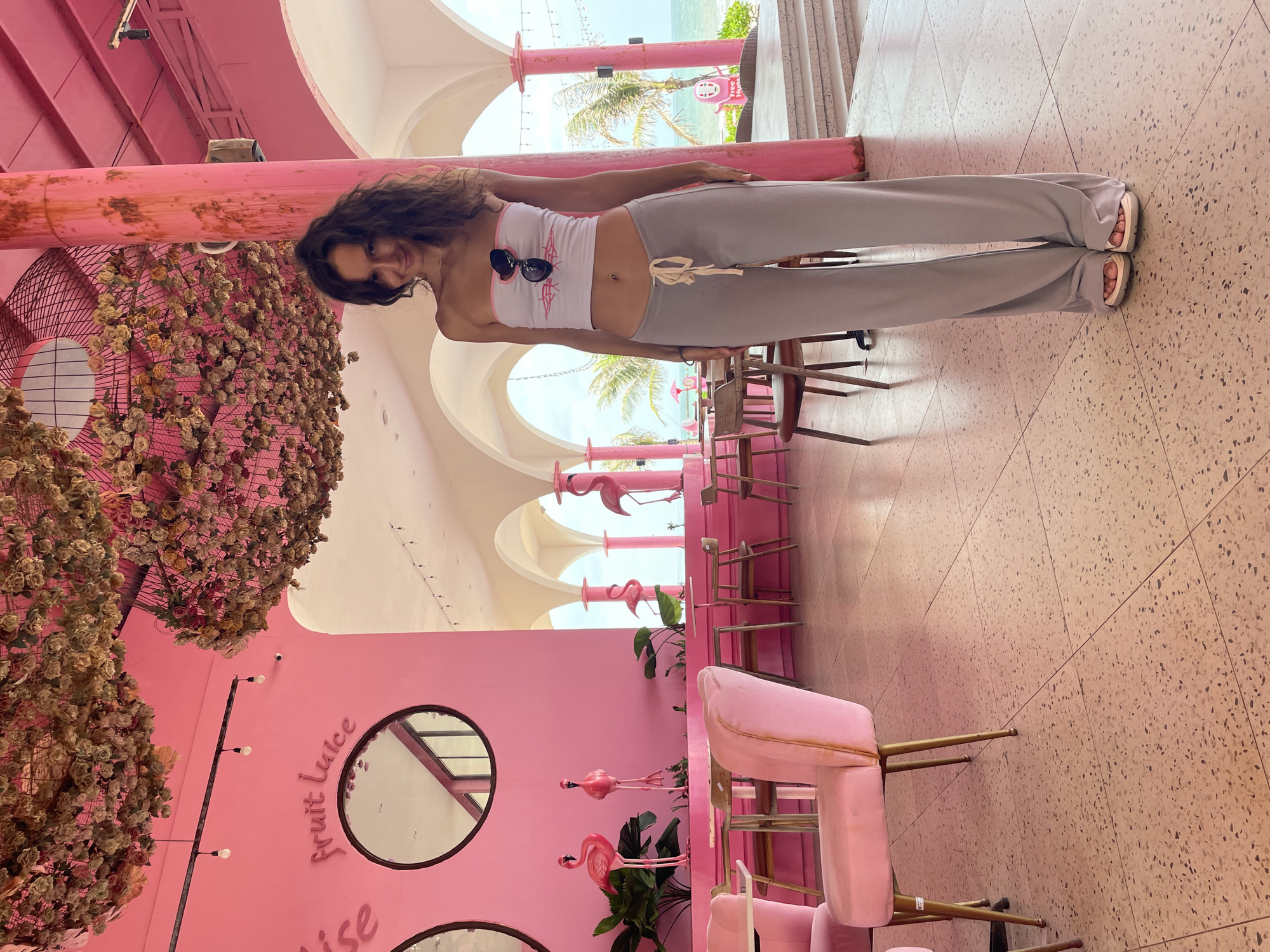The image depicts a woman standing in a brightly colored pink cafe with a view of the ocean. The cafe features pink walls, pink velvet chairs, and flamingo decorations. Suspended from the ceiling are arrangements of dried flowers. The woman is wearing a white tube top with pink trim and light gray wide-leg pants. She has dark curly hair and is smiling at the camera. She is wearing sandals. In the background, the ocean and palm trees are visible through arched openings. The floor is a terrazzo tile in shades of pink and brown. The location is identified as Tien Thanh Ward, Vietnam.