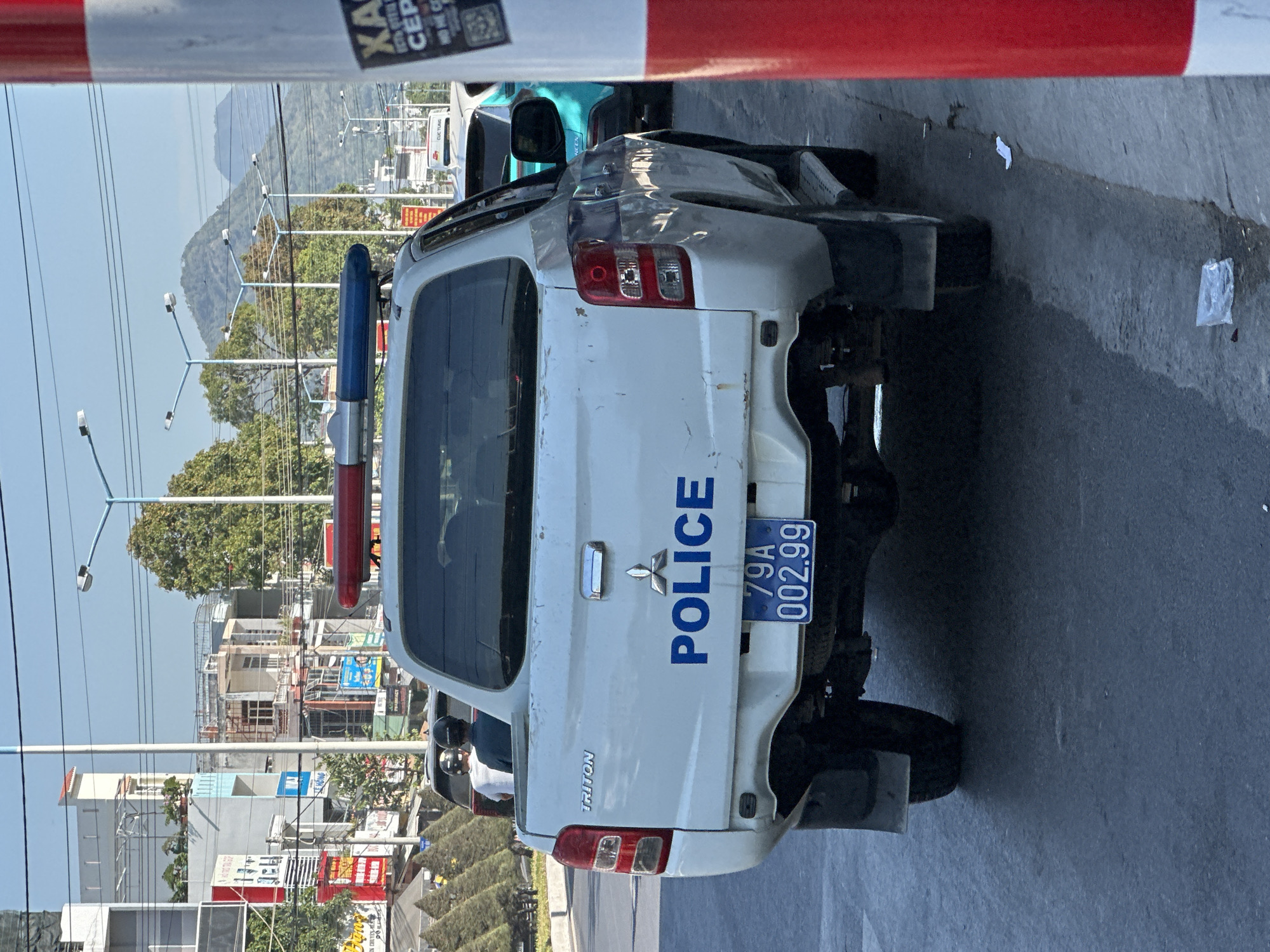 A white Mitsubishi Triton police vehicle is parked on the street in Bac Nha Trang Ward, Vietnam. The rear of the vehicle is severely damaged. The license plate reads "79A 002.99". A blue and red emergency light bar is mounted on the roof. Buildings are visible in the background.