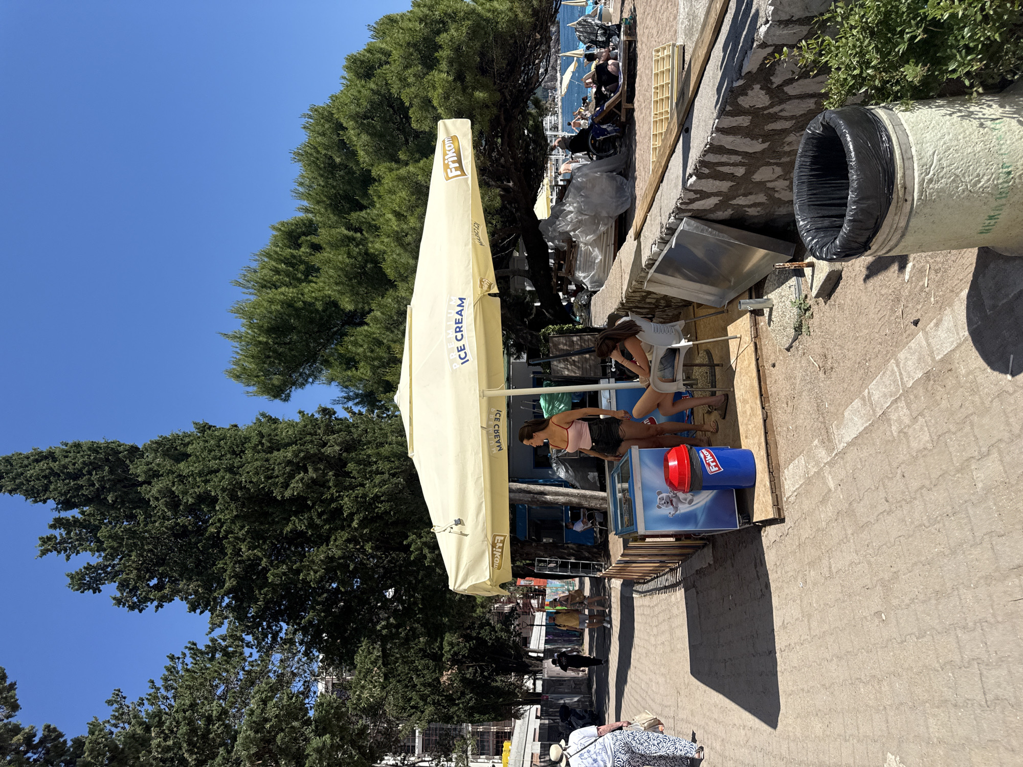 This image depicts a sunlit outdoor scene in Rafailovići, Montenegro, focusing on a pedestrian pathway adjacent to a beach. In the foreground, a light-colored concrete trash receptacle with a black liner is visible on the right, next to a small green bush. The pedestrian path, made of interlocking pavers, extends from the left foreground towards the background, exhibiting strong shadows cast by unseen objects to the left.
In the midground, slightly to the right of the path, a yellow "PREMIUM ICE CREAM" umbrella, branded with "Frikon" on the side, provides shade over a small ice cream stand. Two women are present near the stand: one with long dark hair, wearing a pink tank top and dark shorts, stands barefoot facing left next to an ice cream freezer. The freezer features a graphic of a polar bear. Adjacent to it is a blue trash can with a red lid, also branded "Frikon." The second woman, with light brown hair, is seated on a white plastic chair to the right of the standing woman, facing away from the viewer.
Behind the ice cream stand and to its right, a stone wall separates the path area from the sandy beach. Various materials, including white plastic-wrapped bundles and wooden panels, are stacked along and behind this wall. On the beach, several individuals are visible lounging on sunbeds and under beach umbrellas, with the blue sea and distant coastline forming the horizon.
To the left of the ice cream stand, the paved path continues into the background. A woman in a white patterned top and pants walks away from the viewer on the left side of the path. Further back, several other pedestrians are visible on the path, walking between dense green trees. Some buildings, possibly under construction or renovation, are visible through the trees on the far left. The background is dominated by a clear, bright blue sky above a canopy of dark green trees, including tall, slender cypress-like trees and broader-canopied pines. The overall scene indicates a busy commercial area near a beach on a clear day.