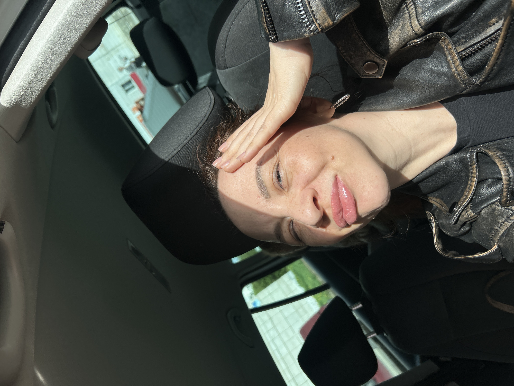 A woman is seated inside a car, possibly a passenger, with her hand resting on her forehead. She is wearing a black leather jacket and a black shirt. The car appears to be parked, as outside the window, buildings and greenery are visible, suggesting an urban environment. The location is Chaikovsky, Russia.