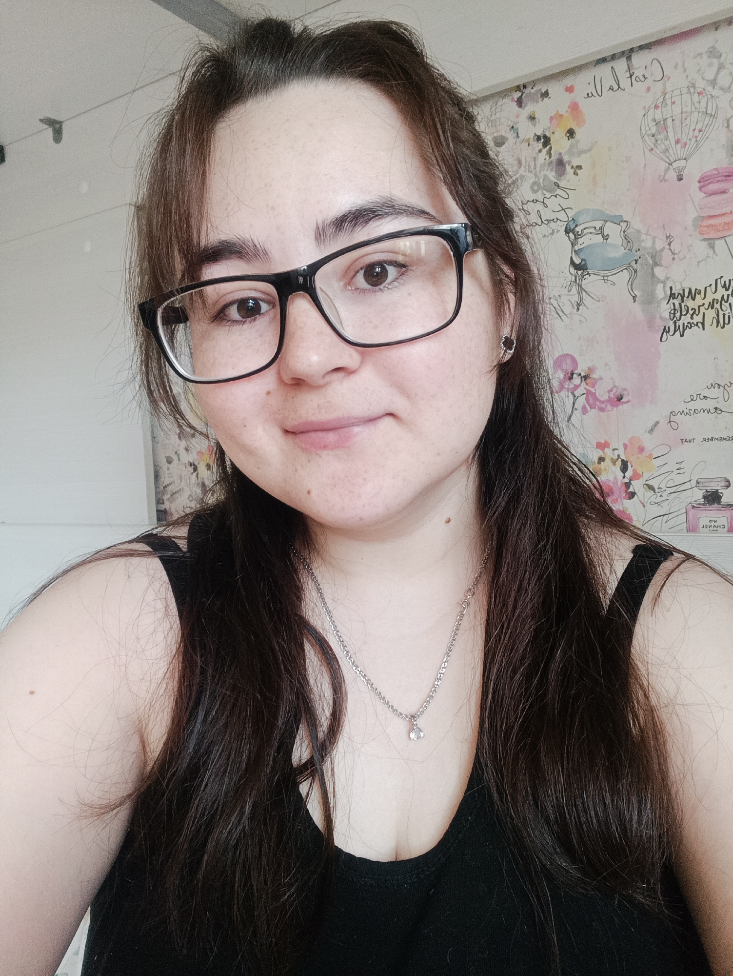 A woman with dark hair, brown eyes, and freckles is wearing black-rimmed glasses and a black tank top. She is wearing a silver necklace. She is in a room with white walls. The wall behind her has a floral and hot air balloon pattern. The city of Noviy, Russia is not visible in the image.