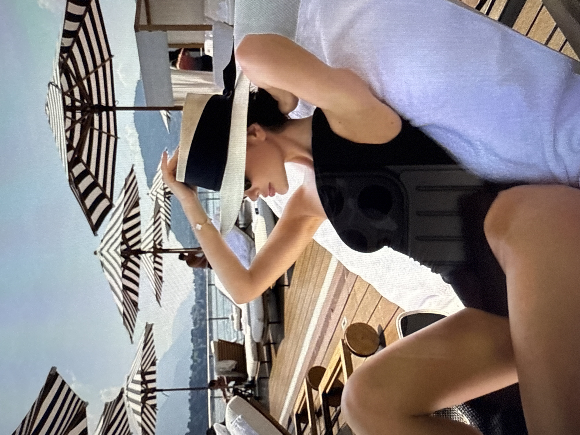 In the city of Găvănoasa, Moldova, a woman is pictured lounging on a deck chair in a resort setting, likely by a body of water. She is wearing a black swimsuit and a wide-brimmed straw hat with a black ribbon. Her left arm is raised, resting on the back of the chair, and her right arm is bent behind her head. The deck is made of wooden planks, and several other deck chairs are visible, some occupied by other guests. In the background, striped umbrellas provide shade, and a body of water with distant hills and mountains can be seen under a clear blue sky. The overall scene suggests a relaxed vacation or resort atmosphere.