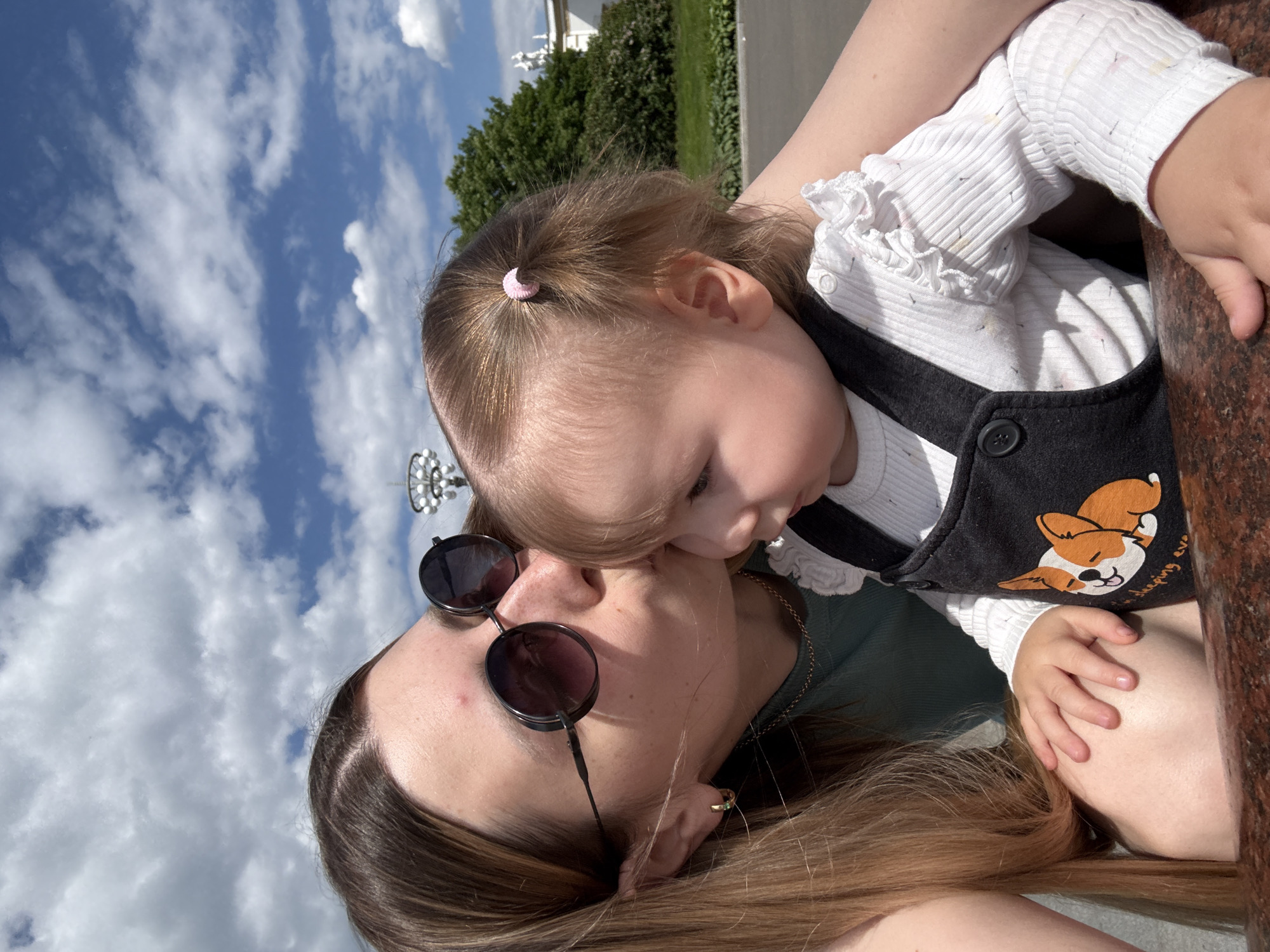 This intimate outdoor portrait shows an adult and a young child sharing a tender moment. The image is rotated 90 degrees clockwise, resulting in the sky appearing on the left side of the frame.
The adult, positioned on the lower right of the image, has long, light brown hair and wears dark, round sunglasses, a green top, and a gold chain necklace. A gold ring is visible on her finger. She is gently kissing or nuzzling the cheek of the child, who is nestled close to her.
The child, appearing to be a toddler, has light brown hair tied with a small pink elastic band. They are dressed in a white, ribbed long-sleeved top with ruffled cuffs and a dark grey or black overall. The overall features a graphic of a light brown and white cartoon corgi dog, with partially obscured text underneath that appears to read "sleeping ever...". The child's face is partly visible, looking downwards.
The background, predominantly blue sky with scattered white clouds, indicates a sunny daytime. Green trees and a distant white architectural structure suggest a park or public area, fitting the context of Moscow, Russia. A reddish-brown polished stone surface, possibly granite or marble, is visible on the right. The image powerfully conveys emotions of affection and warmth between the adult and child.