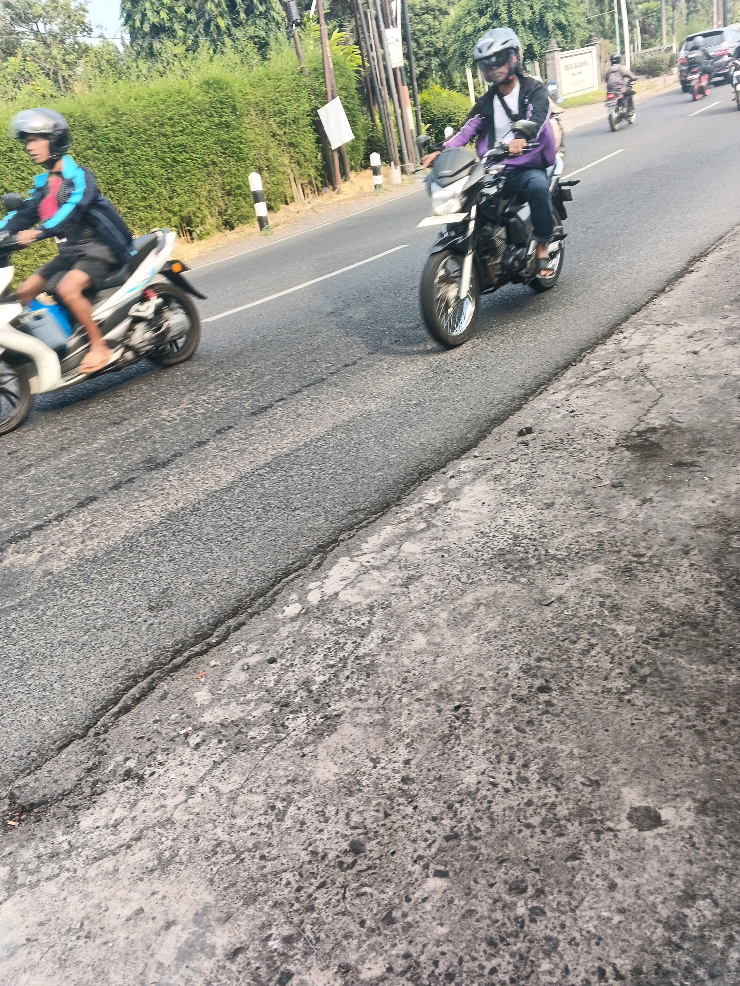 A group of people are riding motorcycles on a paved road. The road has a crack running across it. There are trees and bushes on the side of the road, as well as a white sign on a pole. A man in a purple jacket and jeans is riding a motorcycle, and a person wearing a blue jacket and helmet is on another motorcycle. The scene is set in Ungaran, Indonesia.
