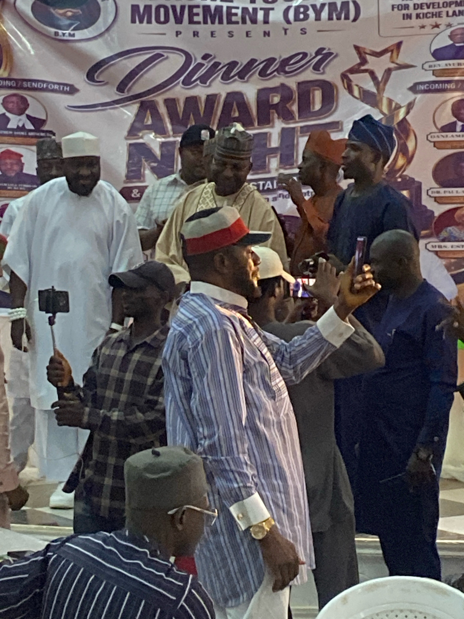 The image captures a group of men in what appears to be an indoor event, likely a ceremony or award presentation, at the O.Y.M Event Center in Jos, Nigeria. In the foreground, a man in a blue and white striped shirt and a dark cap is being photographed. He is wearing a gold watch and has his arm raised, holding what seems to be a phone to take a picture. To his left, another man in a white kaftan and a white cap is also holding a phone, possibly to take a photo or record a video. Further back, several other men are visible, some wearing traditional attire and headwear, including caps and turbans. A large banner with text and graphics, including the words "Dinner Award Night," is prominently displayed in the background. The lighting suggests an indoor event, possibly with artificial lighting. The overall scene depicts a social gathering with elements of celebration and documentation through photography.