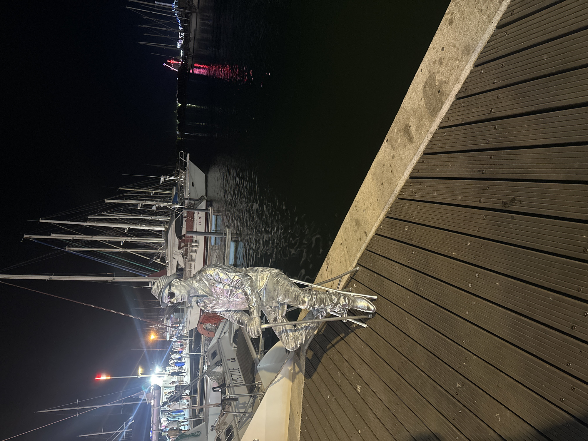 A silver-painted street performer sits on a folding stool on a wooden boardwalk at night in Agde, France, near the marina. The performer is dressed in a shimmering silver outfit, complete with a hat and sunglasses, and holds a silver cane. Behind the performer, several sailboats are moored in the dark water, their masts reaching into the night sky. In the distance, a Ferris wheel with red lights is visible, suggesting a fair or festival. The boardwalk is made of dark wooden planks, and a concrete barrier separates it from the water. Several other boats and a lamppost are also visible in the background, along with a crowd of people in the distance.