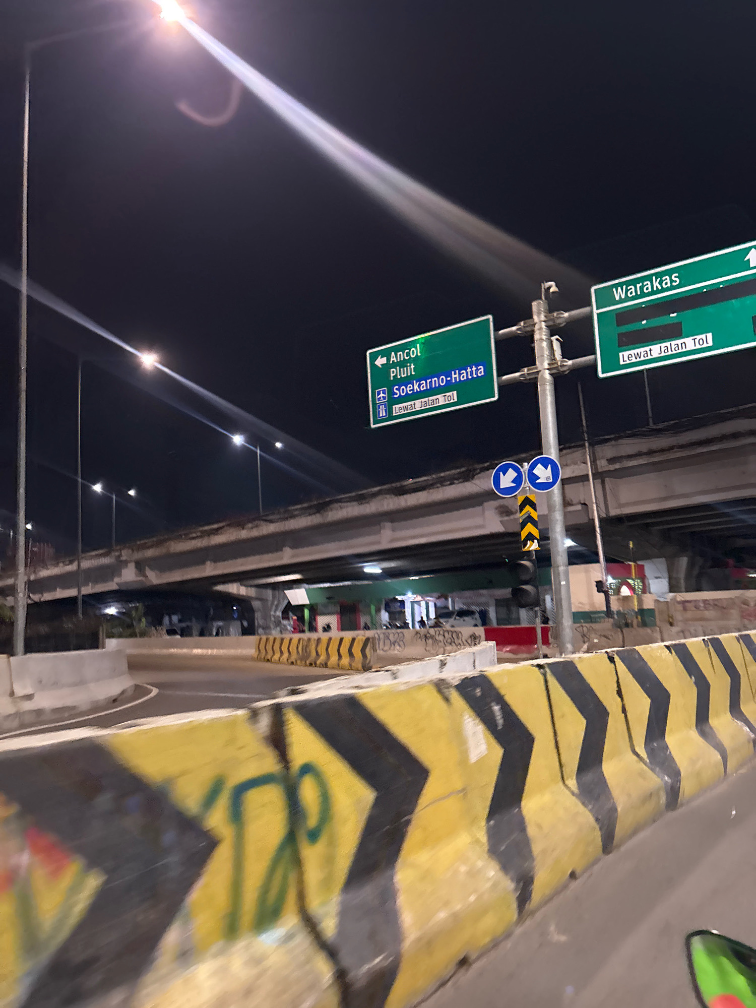 The image depicts a nighttime view of an elevated highway interchange in North Jakarta, Indonesia. The foreground shows a yellow and black striped barrier with graffiti. In the middle ground, traffic signs indicate directions to "Ancol Pluit Soekarno-Hatta" and "Waraka" with options to take the toll road. Blue directional arrows are visible on a pole. The background features an elevated highway structure with streetlights illuminating the scene. The environment is urban, characterized by concrete infrastructure and artificial lighting. No people are clearly identifiable in the image.