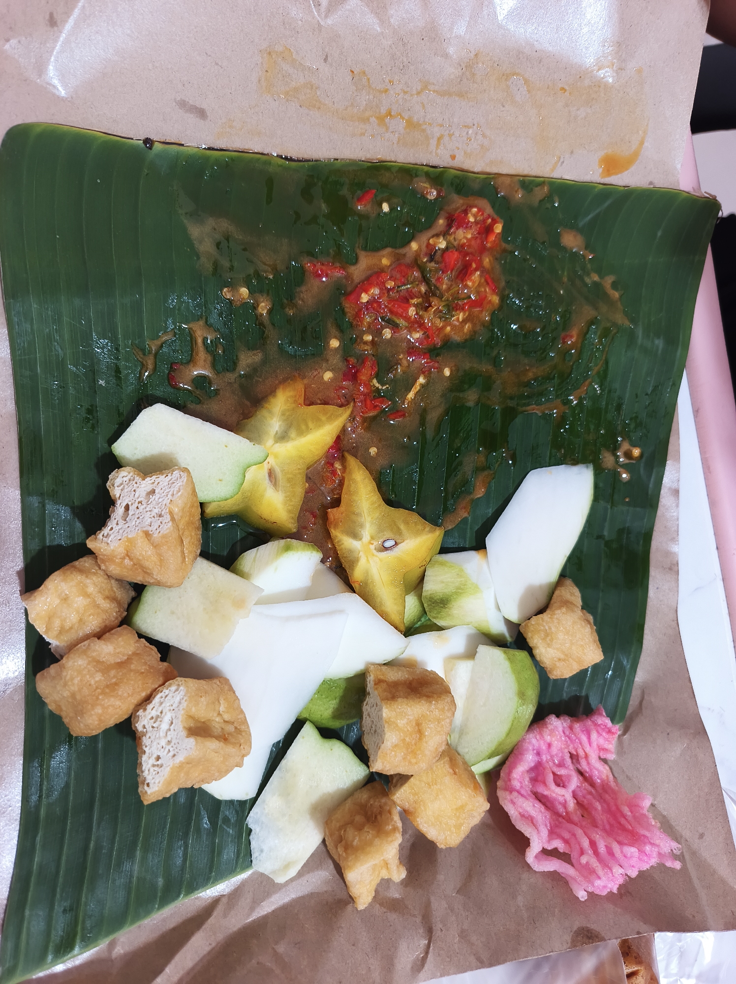 This image captures a close-up, overhead view of a plate of Indonesian fruit and tofu salad, likely served in Jember Kidul, Indonesia. The food is arranged on a large green banana leaf, which itself is placed on brown paper. The salad consists of various fruits, including slices of starfruit and what appears to be unripe mango or guava, cut into wedges. Interspersed with the fruit are cubes of fried tofu. A vibrant pink, crunchy snack, possibly kerupuk, is situated to the right of the main salad. The top portion of the banana leaf is covered in a reddish-brown sauce, visibly containing chopped chilies and seeds, indicating a spicy dressing. The overall impression is of a fresh, savory-sweet dish, typical of Indonesian street food or casual dining. The setting suggests an informal meal, possibly taken outdoors or in a simple eatery.