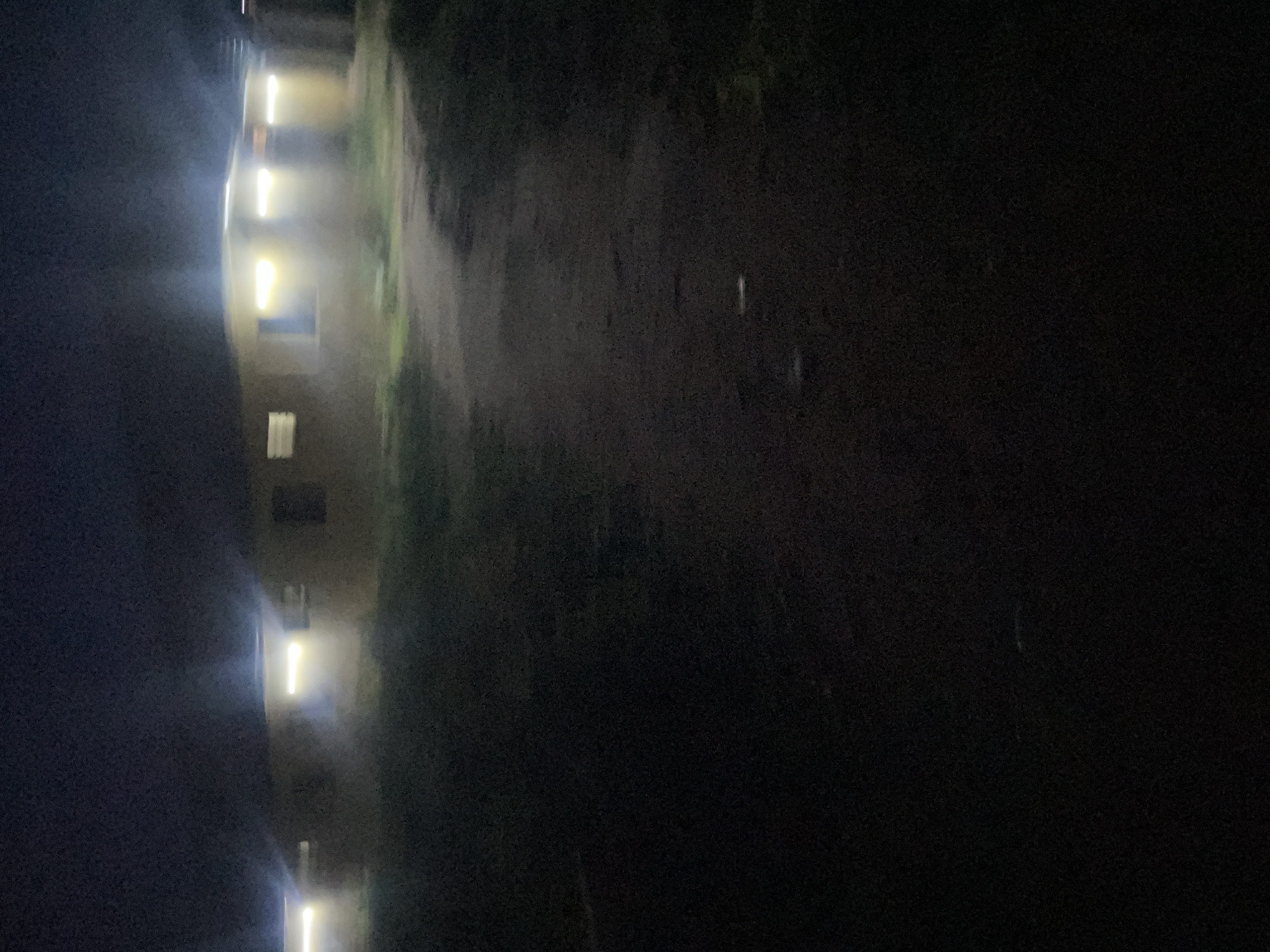 The image depicts a dark, outdoor scene at night, likely in Safari, Nigeria. In the background, several single-story structures are visible, illuminated by bright interior lights shining through windows and doorways. The buildings appear to be made of a light-colored material, possibly concrete or stucco, and are arranged in a row or cluster. A dirt path or road leads towards these structures from the foreground. The ground in the foreground is dark and appears to be unpaved, with some patches of grass or vegetation visible. The overall impression is of a residential or communal area at night, with artificial lighting providing the primary illumination. There are no people visible in the image.