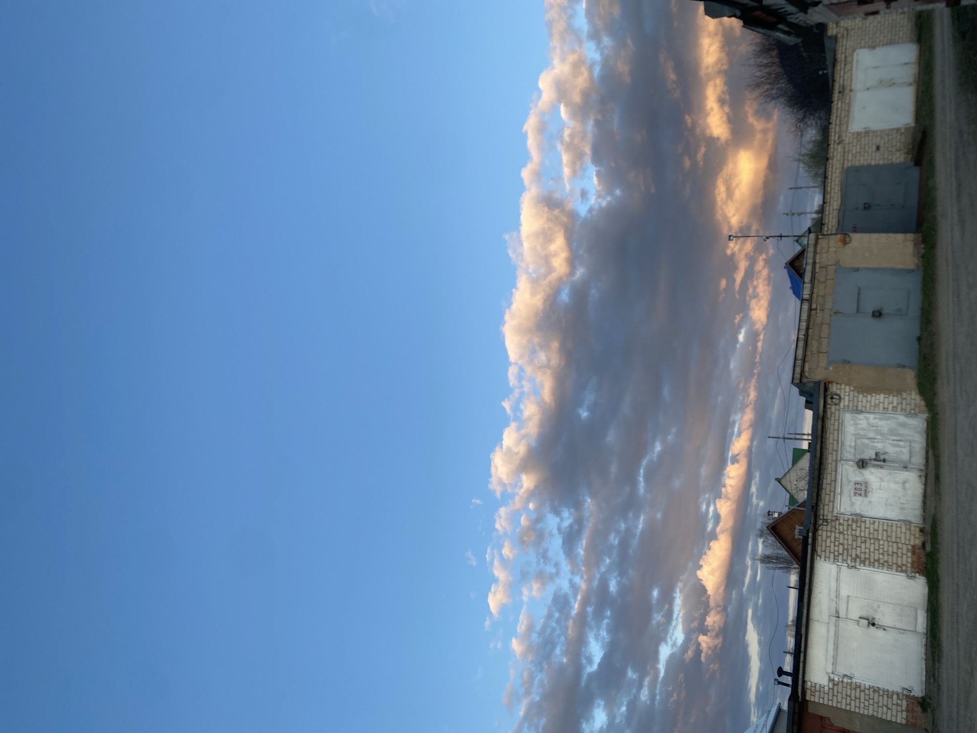 The media file, taken in Chaikovsky, Russia, depicts a serene outdoor scene, likely during either sunrise or sunset. The image is composed of two main sections: a vast expanse of clear blue sky on the left, and a dramatic cloud formation stretching across the right.
The sky transitions from a vibrant, cloudless blue on the far left to a dense band of clouds that extends downwards, almost touching the horizon line where structures begin. These clouds are remarkably textured, with their edges and upper portions illuminated by a warm, golden-orange light, indicative of either early morning or late afternoon sun. The undersides and deeper parts of the clouds appear in various shades of grey and dark blue, creating a striking contrast with the illuminated sections.
Along the lower right portion of the image, a row of garages is visible. These structures are built from light-colored bricks or cinder blocks. Each garage features a metal door; some are painted white, while others are a dull grey. On one of the white garage doors, the number "203" is distinctly painted in dark digits. Behind the garages, the silhouettes of bare trees suggest a season without foliage, possibly winter or early spring. Utility lines or power lines are discernible strung across the scene above the garages. A dirt or gravel path runs along the ground in front of the garages.
No specific activity or event is captured, presenting a still life of an urban or semi-urban landscape. The time of day, marked by the distinctive golden light on the clouds, lends a quiet and atmospheric quality to the scene. The weather appears calm and clear, with no signs of precipitation.