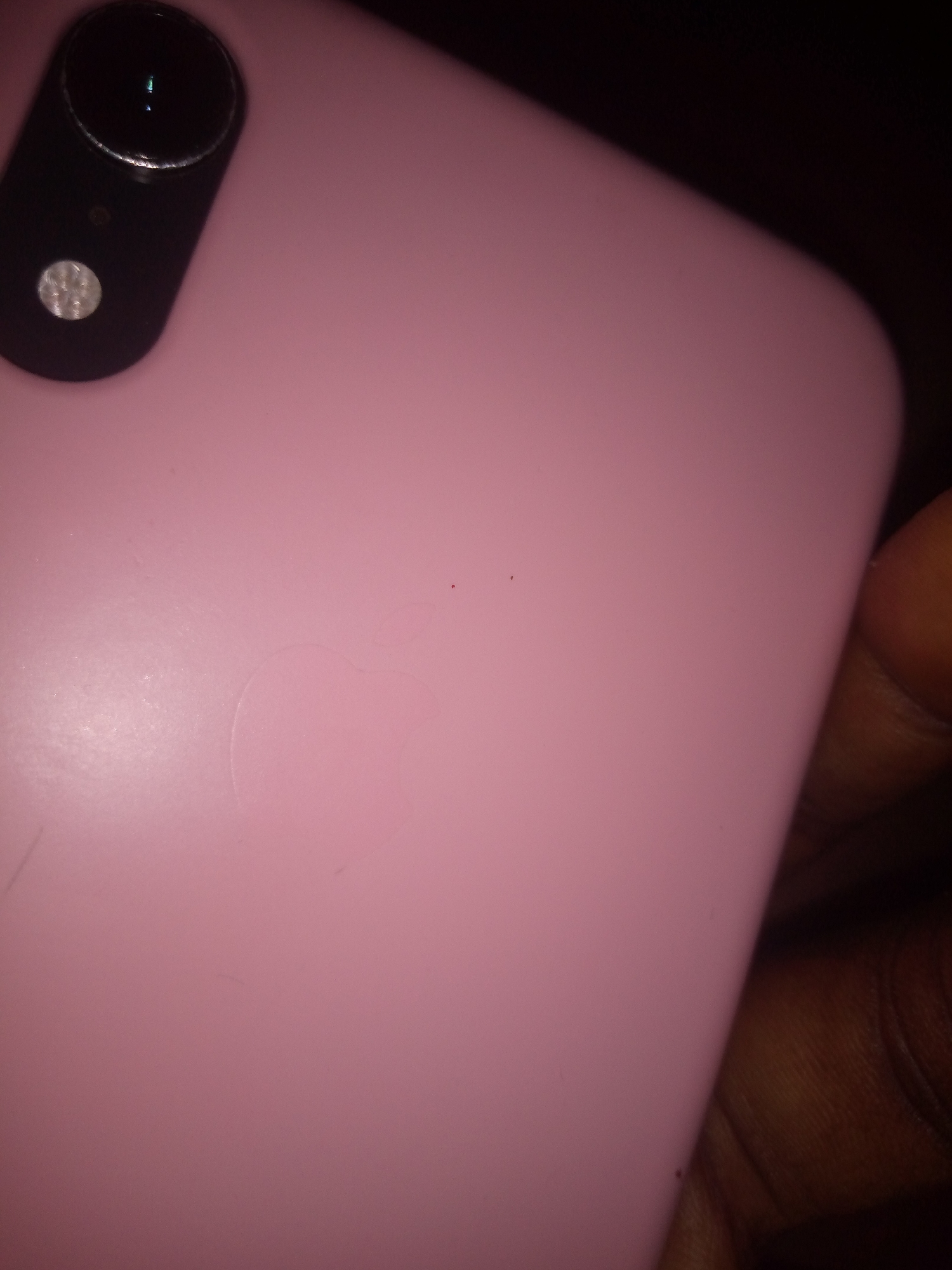 A pink iPhone is lying on its back, facing upwards. The phone has a visible Apple logo etched into the back. The camera lens is in the upper left corner of the phone. There is a small black dot on the phone's surface, located in the upper middle section of the phone. A thumb is visible on the right side of the image, holding the phone. The background is dark and out of focus.
