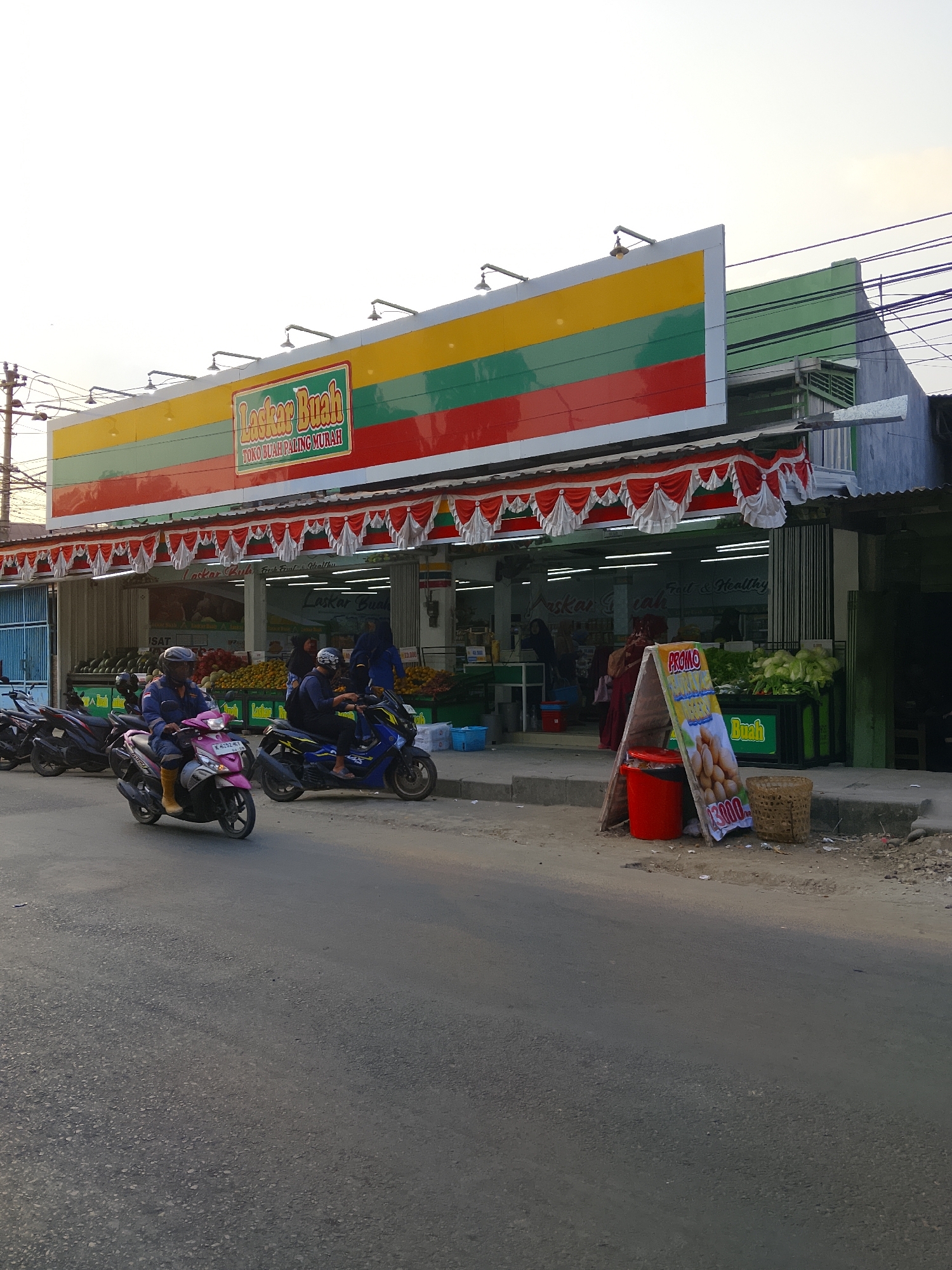 This is a medium shot, eye-level, outdoor, full-color photograph of a fruit shop called "Laskar Buah" located on a street in Karangboyo, Indonesia. The shop has a prominent facade with a large, colorful sign featuring horizontal stripes of yellow, green, and red, with the text "Laskar Buah" and "TOKO BUAH PALING MURAH" in red and white lettering. Red and white decorative fabric valances hang along the front of the shop.
Inside the shop, visible through the open entrance, are displays of fruits and vegetables. Several people are seen inside, some of whom appear to be customers or staff. In front of the shop, on the sidewalk, is a sidewalk A-frame sign with the word "PROMO" at the top and an image of potatoes, with the price "13.000" displayed. Next to the sign is a red bucket and a woven basket.
On the street in front of the shop, three motorcycles are parked or moving. Two men are riding motorcycles: one on a pink scooter in the foreground, wearing a blue jacket and yellow boots, and another on a blue scooter to the right, wearing a dark helmet. A third black motorcycle is parked behind the pink scooter. The street is paved with asphalt, and there is a concrete sidewalk along the shop. Electrical wires are visible overhead. The sky is overcast and bright.