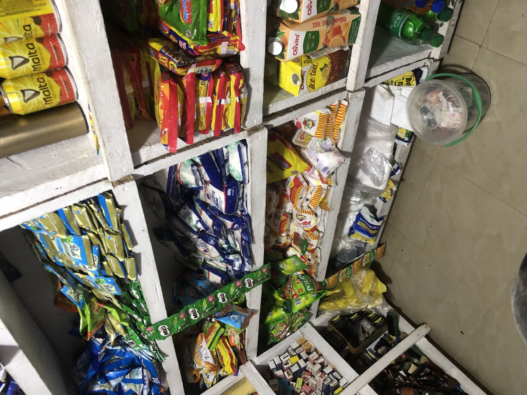 The image depicts a dimly lit interior of a small shop, likely a convenience store or kiosk, with shelves stocked full of various packaged goods. The shelves are made of rough, painted wood, and the overall impression is one of a tightly packed retail space. The scene is set in Ado, Nigeria.
On the top shelf, several cans of "Malta Guinness" are visible, alongside other assorted packaged foods and drinks. The middle shelf displays a large quantity of packaged food items, including what appear to be snacks and possibly instant noodles, stacked in colorful red, yellow, and blue packaging. A string of "Milo" packets hangs down from this shelf. The bottom shelf is also densely populated with various food items, including sachets of "Peak Milk" and packets of sugar. Further down, a large plastic bucket with a green handle and a clear lid sits on the floor, containing various items, possibly money or other small goods. A green bottle of a beverage, possibly a soft drink, is visible on the far right of the bottom shelf. The floor is tiled with neutral-colored tiles. The lighting is artificial and somewhat harsh, casting shadows and highlighting the texture of the shelves and packaging. There are no people visible in the image, and the focus is entirely on the merchandise and the shop's interior.