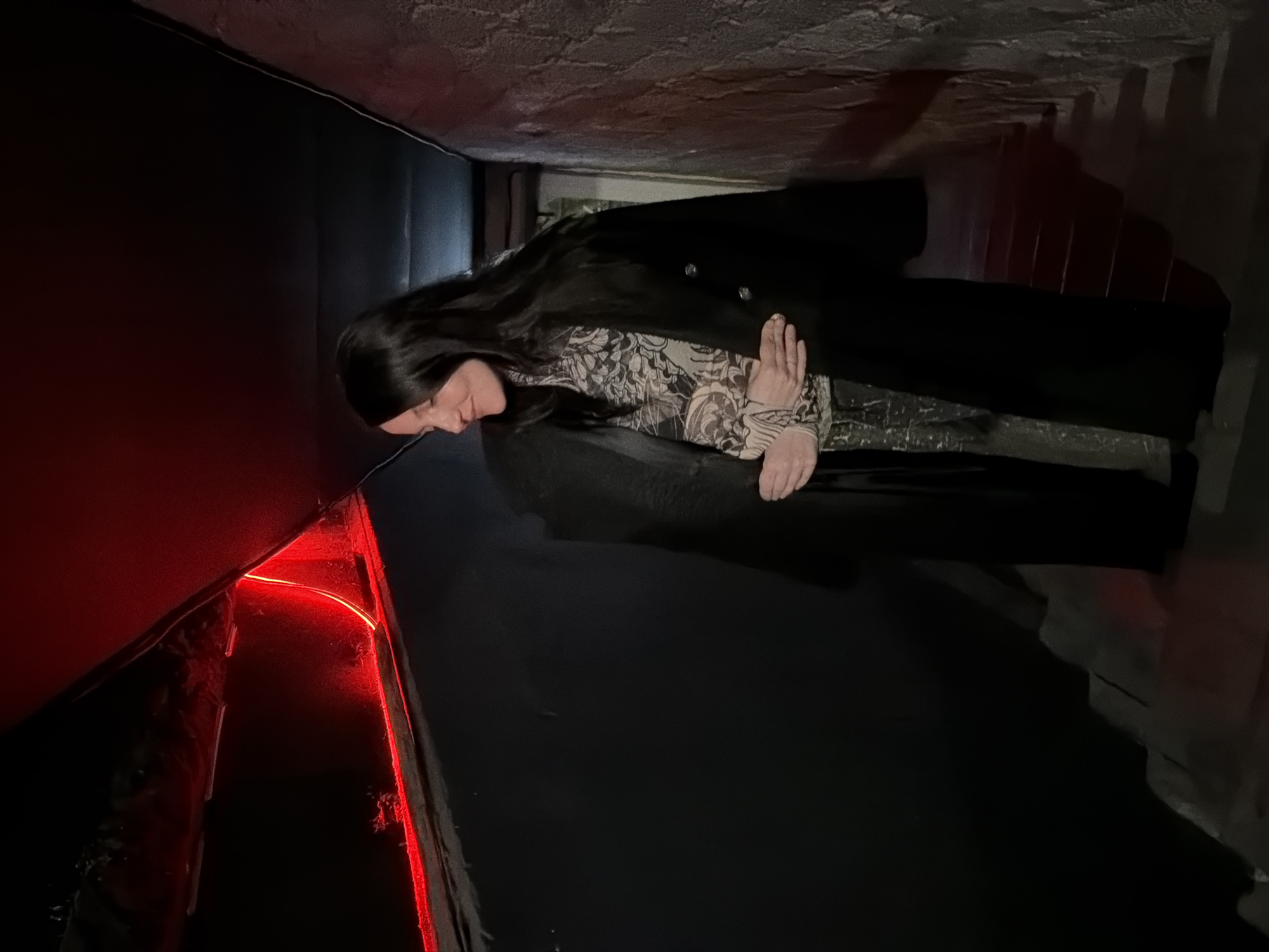 The image shows a woman with long, dark hair wearing a patterned top and a dark coat. She is standing or sitting in a confined space with dark walls, possibly a narrow tunnel or corridor. There is red lighting on one side of the enclosure. The location is Chaikovsky, Russia.
