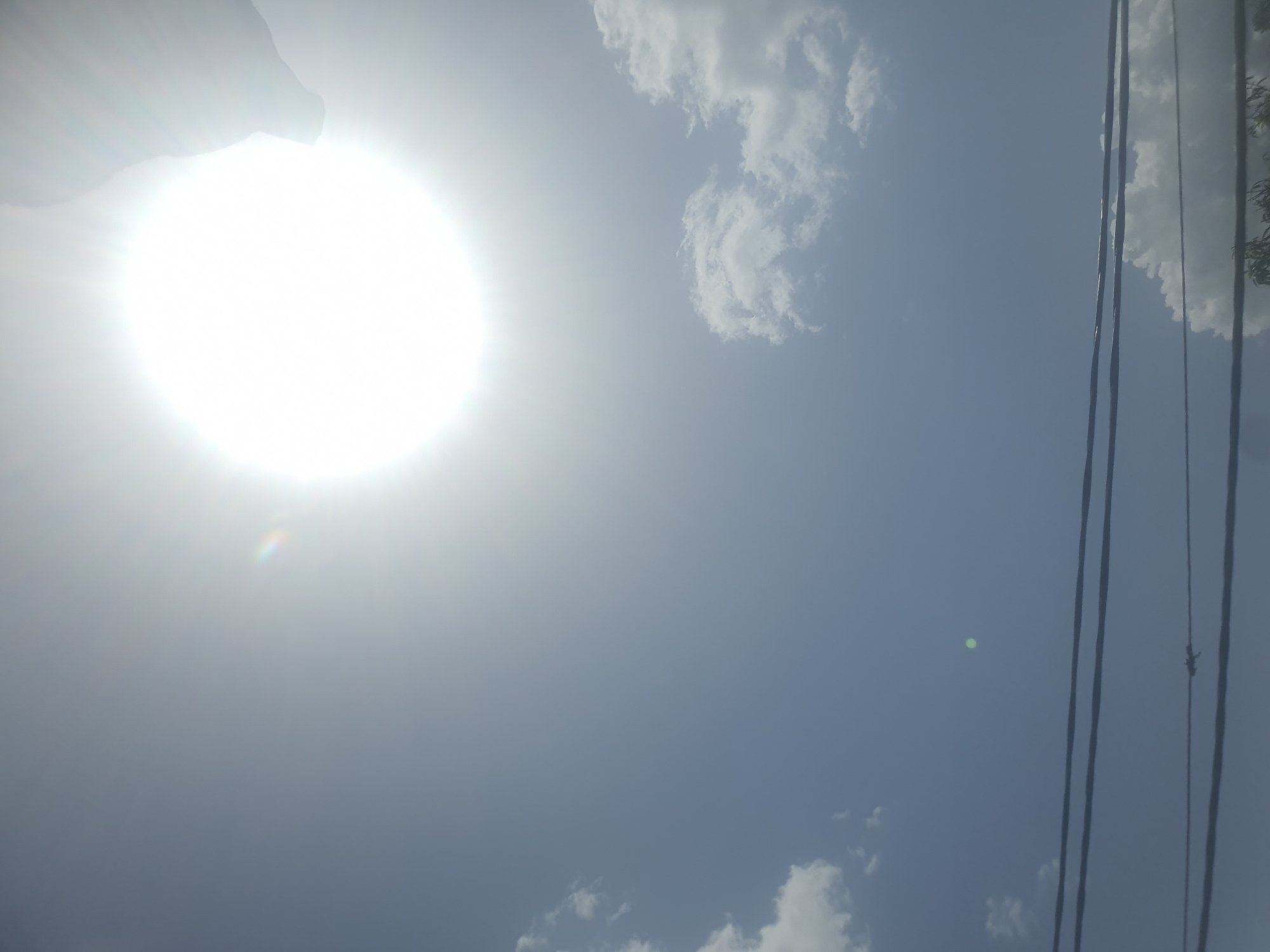 The image depicts a bright, sunny day in Jos, Nigeria. The sun is prominently visible in the upper part of the frame, emitting a strong glare. A few scattered white clouds are present in the sky. In the foreground, several dark, horizontal power lines stretch across the scene. A faint, small green circle is visible in the sky to the left of the power lines. The overall impression is of an outdoor, daytime environment, likely a street or open area with utility infrastructure.