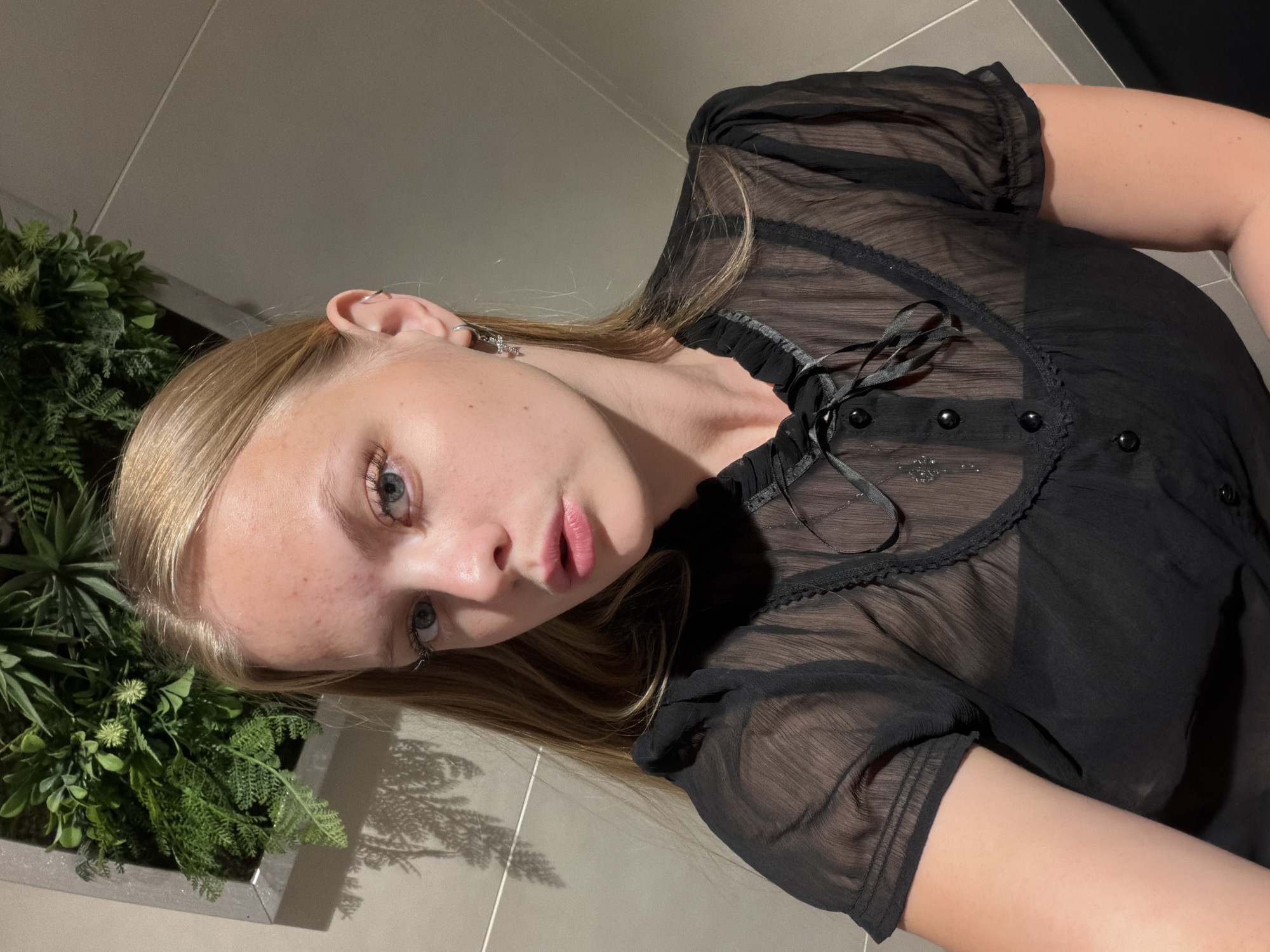 A young woman with blonde hair is lying down, looking up at the camera. She is wearing a sheer black top with ruffles and buttons. She has light blue eyes and pink lips. To her left is a planter filled with greenery, casting a shadow on the tiled floor. The lighting suggests it is indoors, possibly in a bathroom or kitchen, given the tiled walls and floor. The overall mood is casual and relaxed. The image was taken in Izhevsk, Russia.