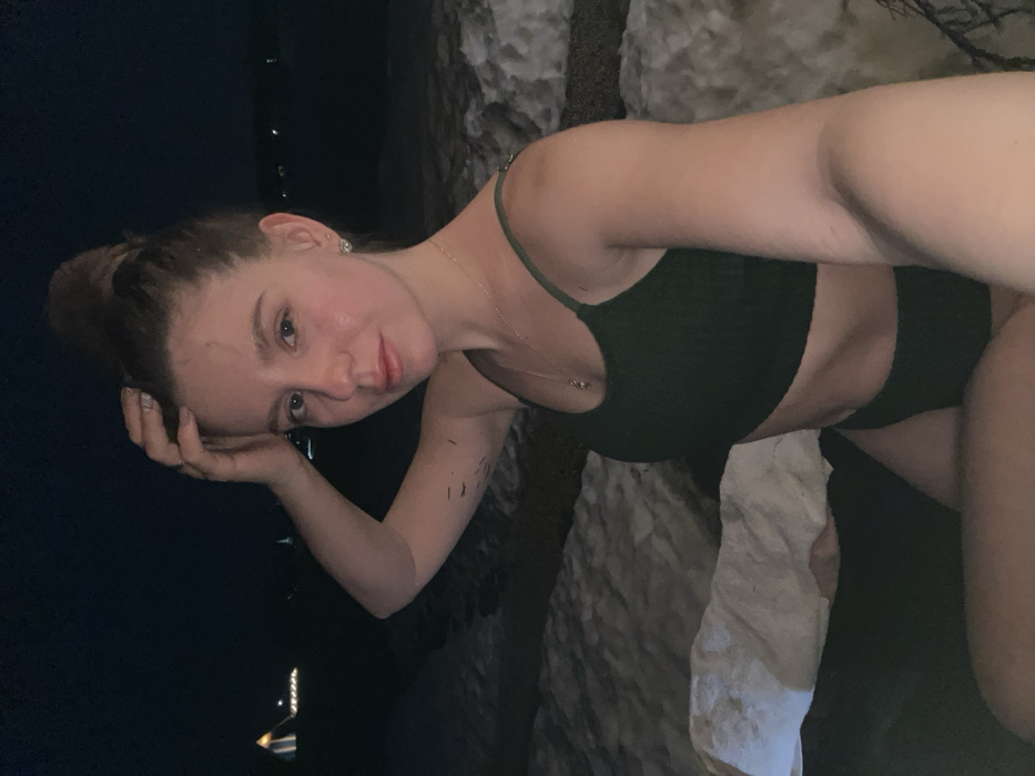 A young adult female is depicted in a close-up, waist-up, selfie-style photograph taken at night in an outdoor, snowy environment. She has light skin, light-colored eyes, and her hair is styled in a high bun. She wears a dark green ribbed sports bra or bralette and visible dark green briefs. A delicate gold-colored chain necklace with a small pendant and stud earrings are visible. Her left hand is raised, fingers touching the hair above her forehead. Multiple thin, dark, linear marks are visible on her left forearm. The background is dark, indicating nighttime, with patches of snow or ice visible on the ground directly behind her. In the far distance, several artificial lights are present, including one structure with an illuminated pyramidal or tent-like shape. The specific location within Shipulino, Russia, is not identifiable from the visual information provided in the image.