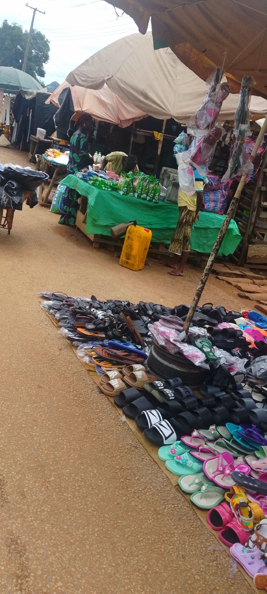 This is a photograph taken in Baissa, Nigeria, depicting a bustling outdoor market scene. The foreground is dominated by a large display of footwear, primarily flip-flops and sandals, spread out on the ground. Various colors and styles are visible, suggesting a diverse selection for customers.
In the midground, several stalls are set up. One stall, covered with a green cloth, displays bottles of what appears to be beverages, along with other packaged goods. Behind this stall, a person in a patterned dress is partially visible. To the left of this stall, another person in a green and black patterned garment stands near a table with items. Further back, clothing items hang on display.
The setting appears to be a roadside market, with a dirt or gravel road surface in the foreground. The sky is overcast, suggesting it might be daytime with cloudy weather. Overhead, a large, light-colored tarp provides shade for some of the stalls, supported by wooden poles. The overall impression is one of a vibrant local marketplace, likely busy with shoppers and vendors. There is no visible text in the image.