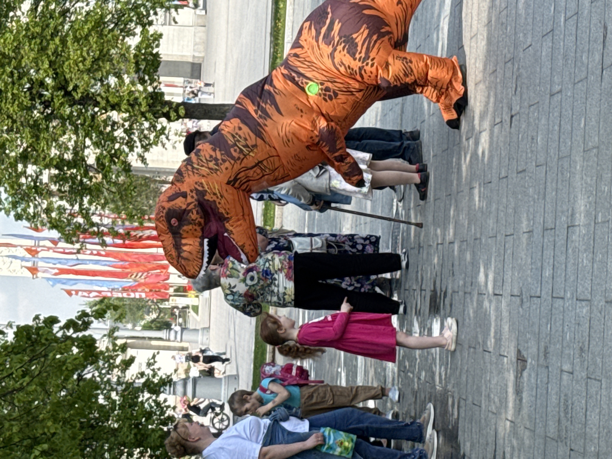 A group of people, including children, stand on a paved surface near a street in Moscow, Russia. One person wears an inflatable orange and brown dinosaur costume. Flags are visible in the background. Trees are present on the sides of the image.