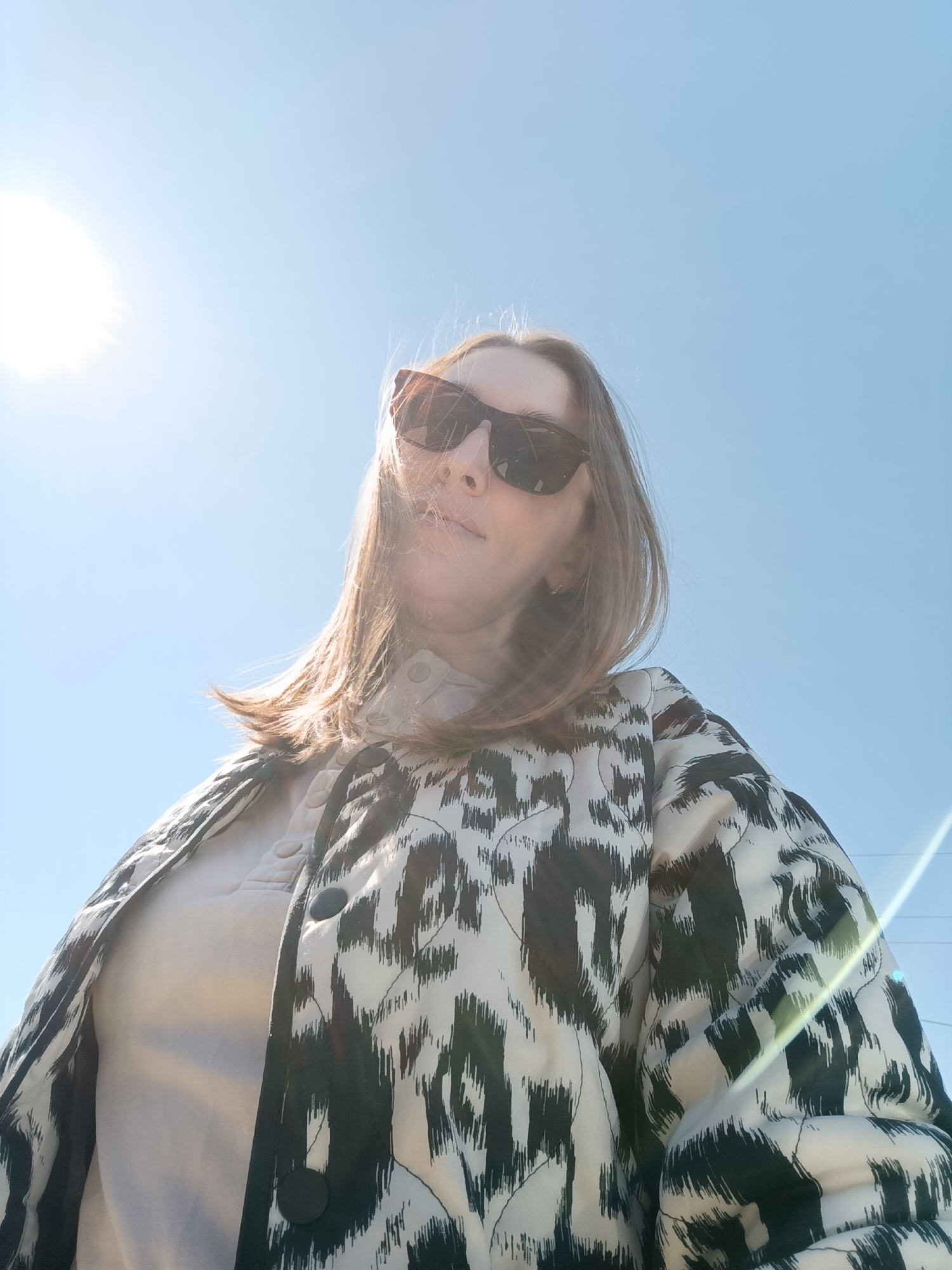 A woman is photographed from a low angle against a bright, clear blue sky with the sun visible in the upper left. She is wearing dark sunglasses and a patterned jacket with black and white abstract designs. A light-colored shirt is visible underneath the jacket. Her hair is light brown. The photograph appears to be taken outdoors, but no specific location is identifiable from the image. The location of Chaikovsky, Russia cannot be verified from the visual information provided.