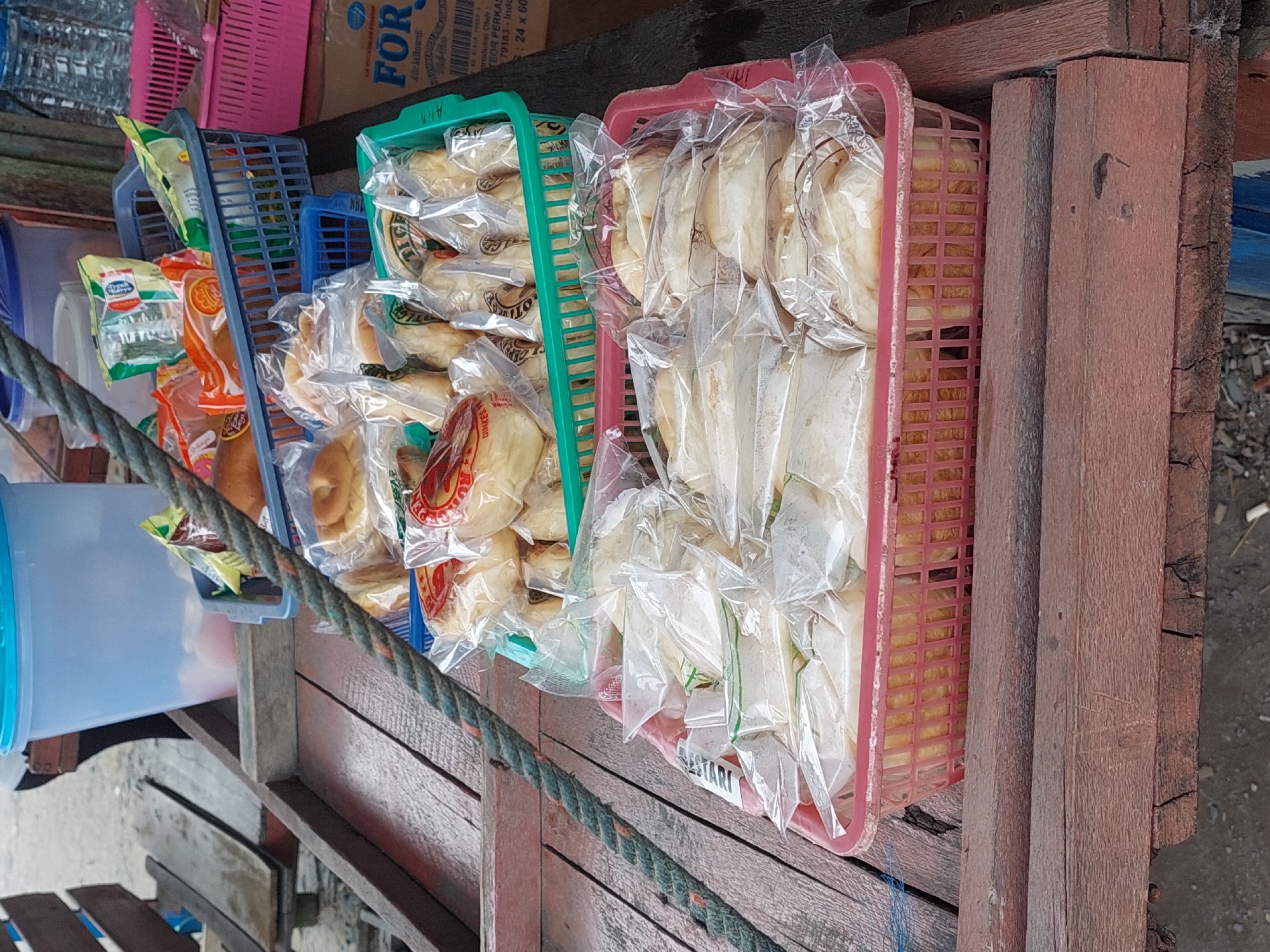 In Parit Baru, Indonesia, a close-up shot captures a rustic wooden market stall overflowing with baked goods. In the foreground, two red plastic baskets are filled with individually wrapped buns. The top basket, a vibrant green, contains more buns also individually wrapped. Behind these, a blue plastic basket holds an assortment of packaged snacks, some with colorful wrappers. In the background, a large blue translucent plastic container is visible, and a pink plastic basket sits on a cardboard box. The overall scene suggests a street-side vendor operation.