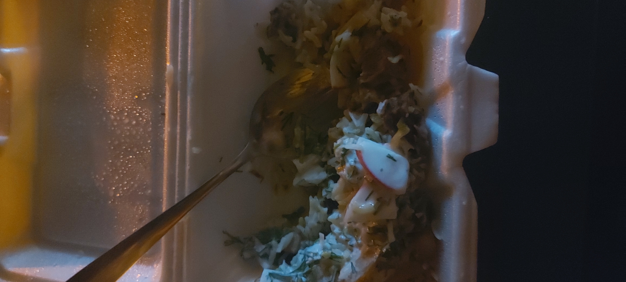 A close-up shot shows a half-eaten meal in a white styrofoam container, possibly taken outdoors at night. The meal appears to be rice with meat and what might be sliced radishes or similar vegetables. A metal spoon rests in the food. The location is unknown; there is no clear indication that the image was taken in Tashkent, Uzbekistan. The lighting is poor and grainy.