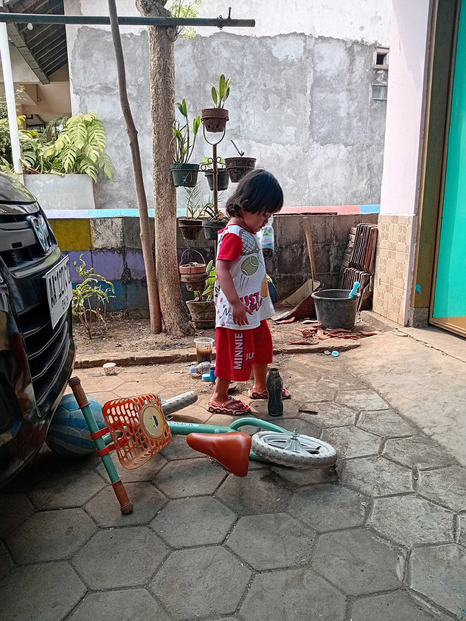 In Gebang, Indonesia, a young child stands next to a partially disassembled bicycle on a paved courtyard. The child, wearing a red and white t-shirt with images of sports equipment and red shorts with "MINNIE" printed on them, looks down at the bicycle. The bicycle, lying on its side, has a detached wheel and a detached orange basket. A black car is parked to the left of the scene, with its front bumper and license plate visible. In the background, a weathered wall with colorful painted sections, several potted plants, and a large gray bucket are present. The courtyard is paved with hexagonal concrete tiles. The setting appears to be a residential outdoor area.