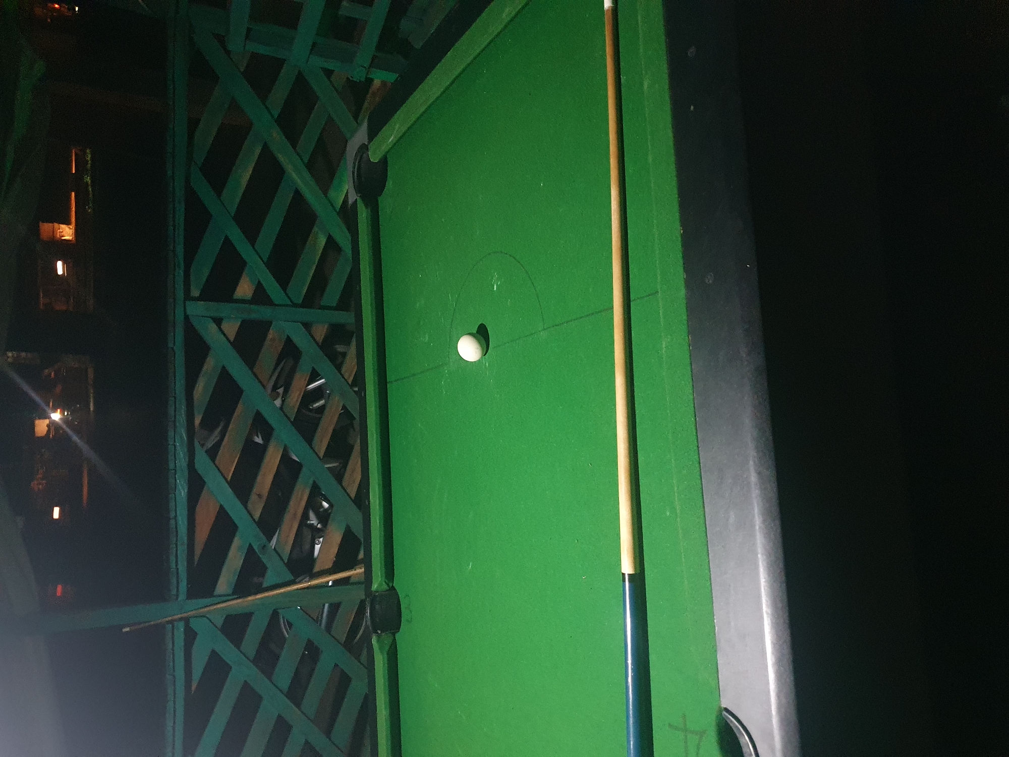 An outdoor pool table with green felt is shown at night, positioned under a covered structure with a green lattice fence. A single white billiard ball rests near the center of the table. A pool cue, with a blue and wood-colored shaft, is placed horizontally along the bottom edge of the table. In the background, through the lattice fence, glimpses of what appear to be bicycles and possibly other objects are visible. The scene is illuminated by artificial light, casting a dim glow. The location is specified as Lokoja, Nigeria.